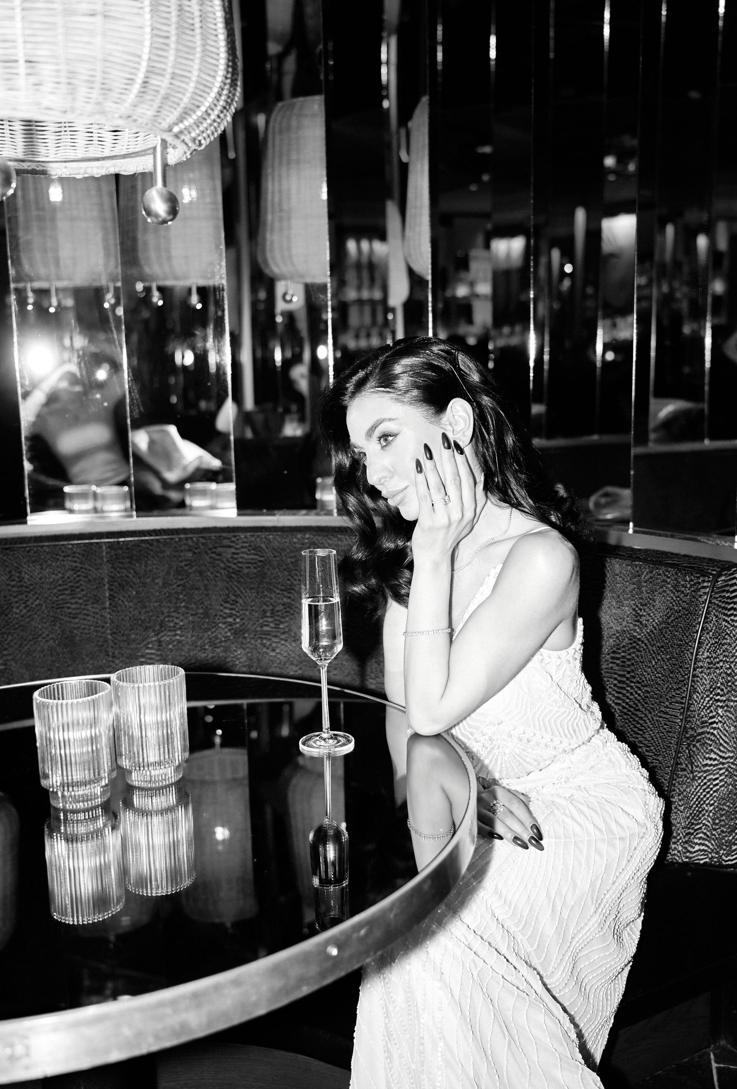 Black and white photo of elegant woman in white dress sitting at bar table with champagne flute.