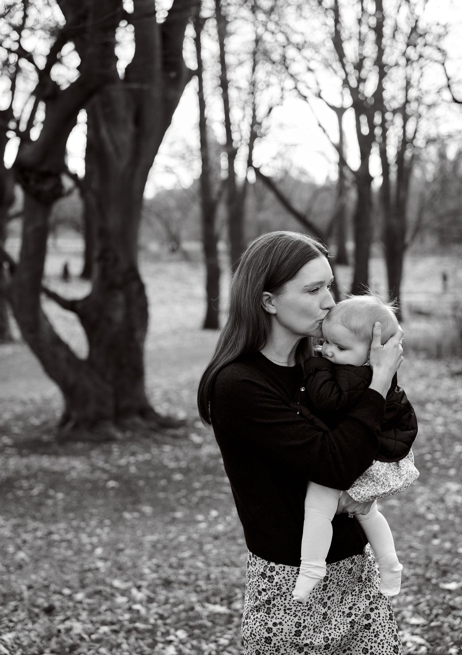 Black and white photo of a mother kissing her baby in a park surrounded by bare trees.