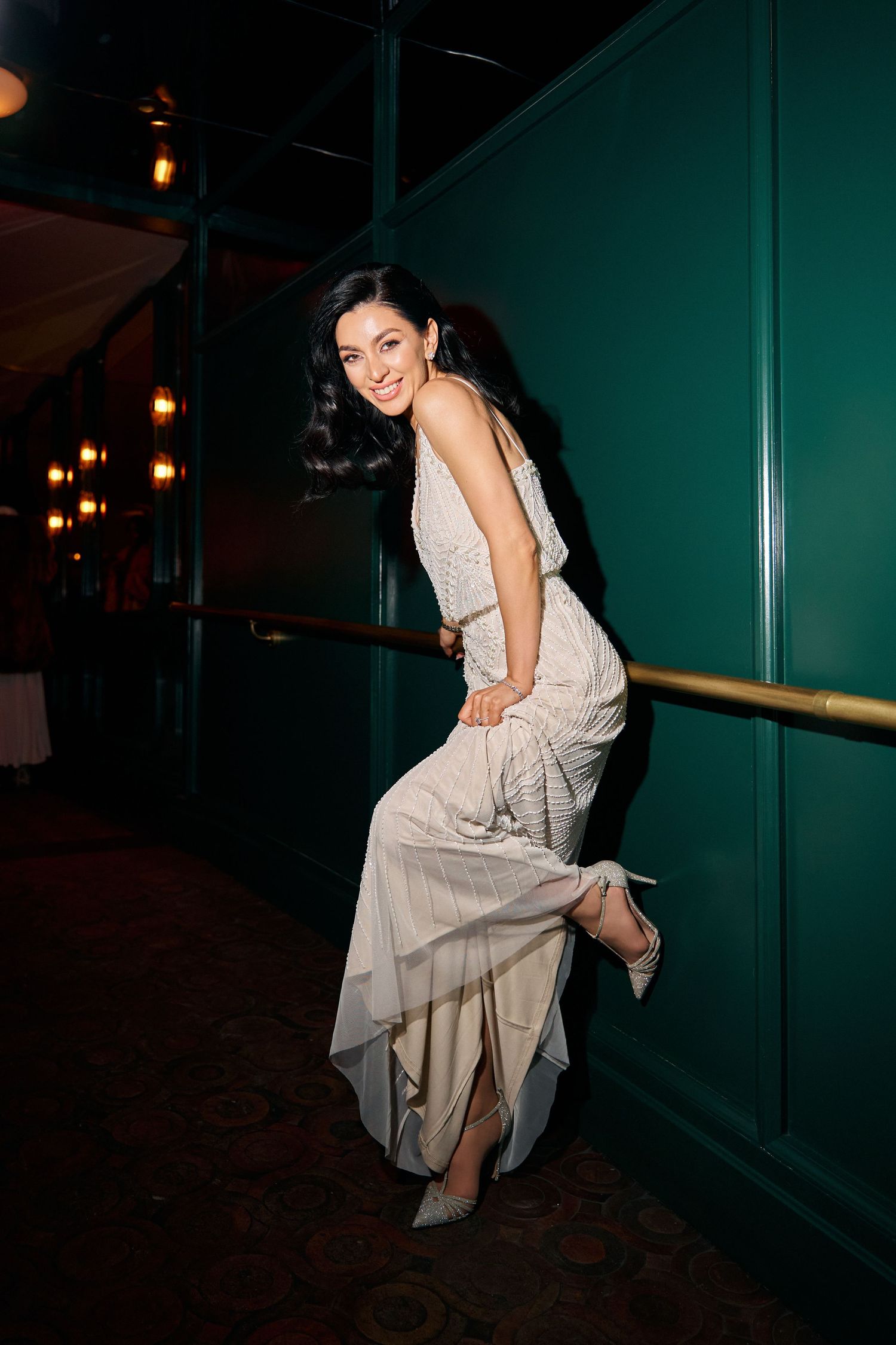 Elegant woman in a beaded ivory gown smiling and leaning against a railing in a dimly lit venue.