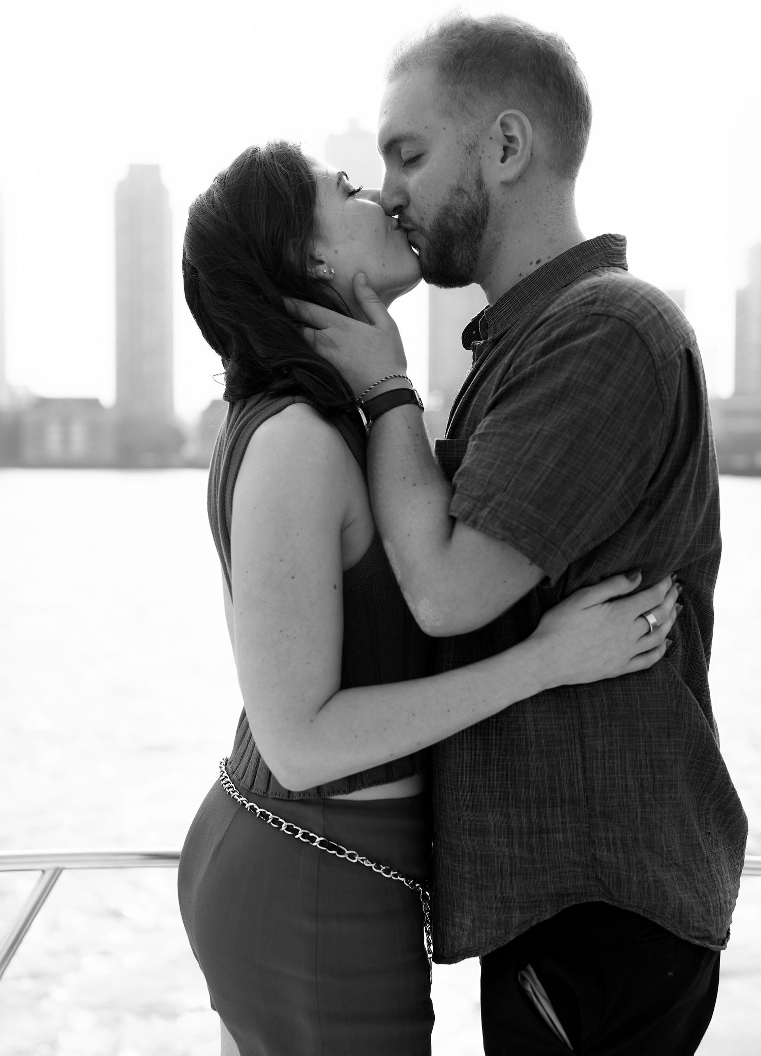 Black and white photo of couple sharing a romantic kiss on a boat with city skyline background.