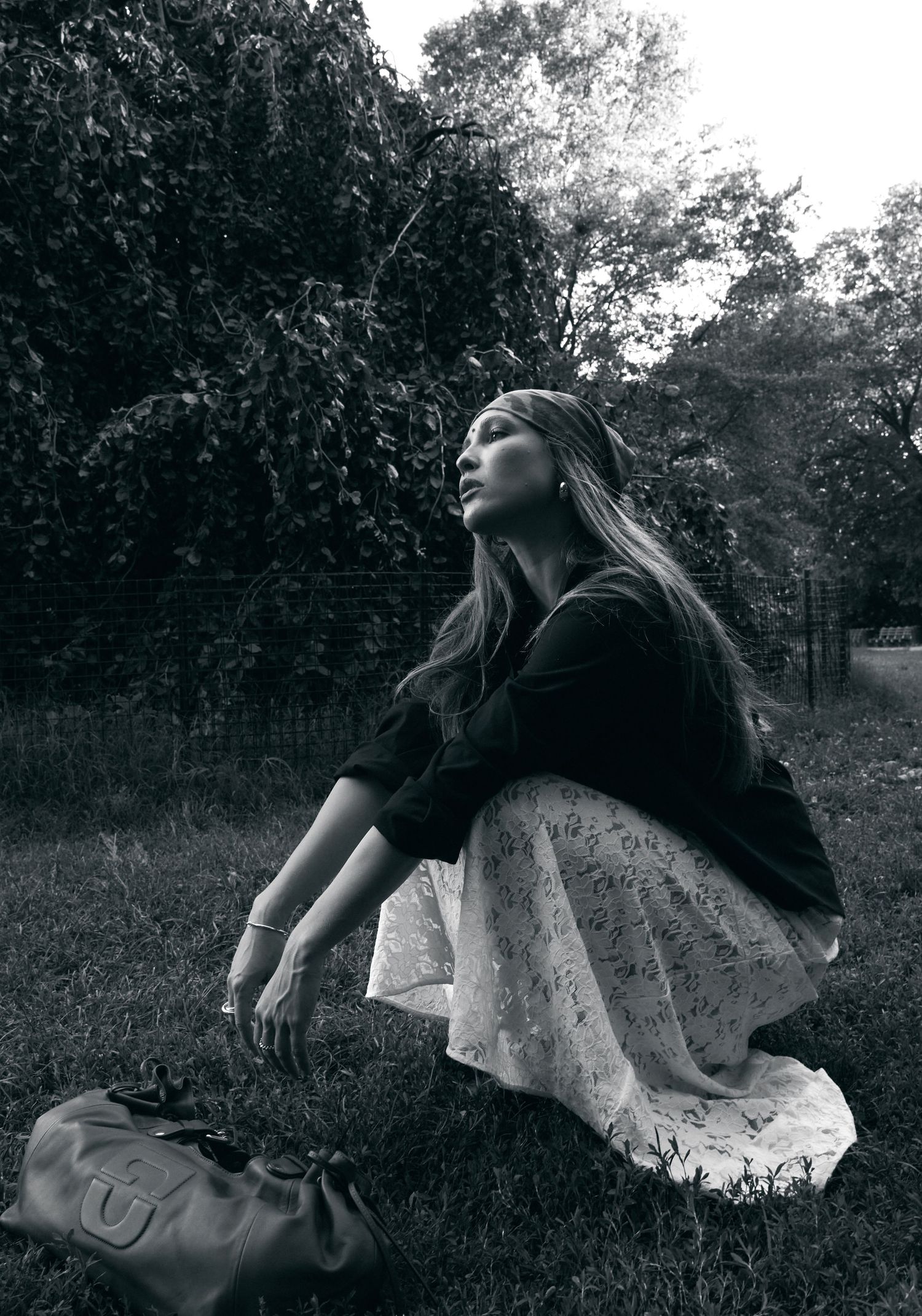 Black and white portrait of woman in lace skirt sitting on grass looking upward near leather bag.