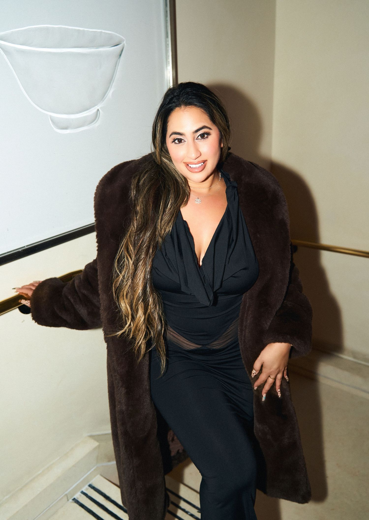 Woman in black dress and brown fur coat poses on staircase with long highlighted hair.