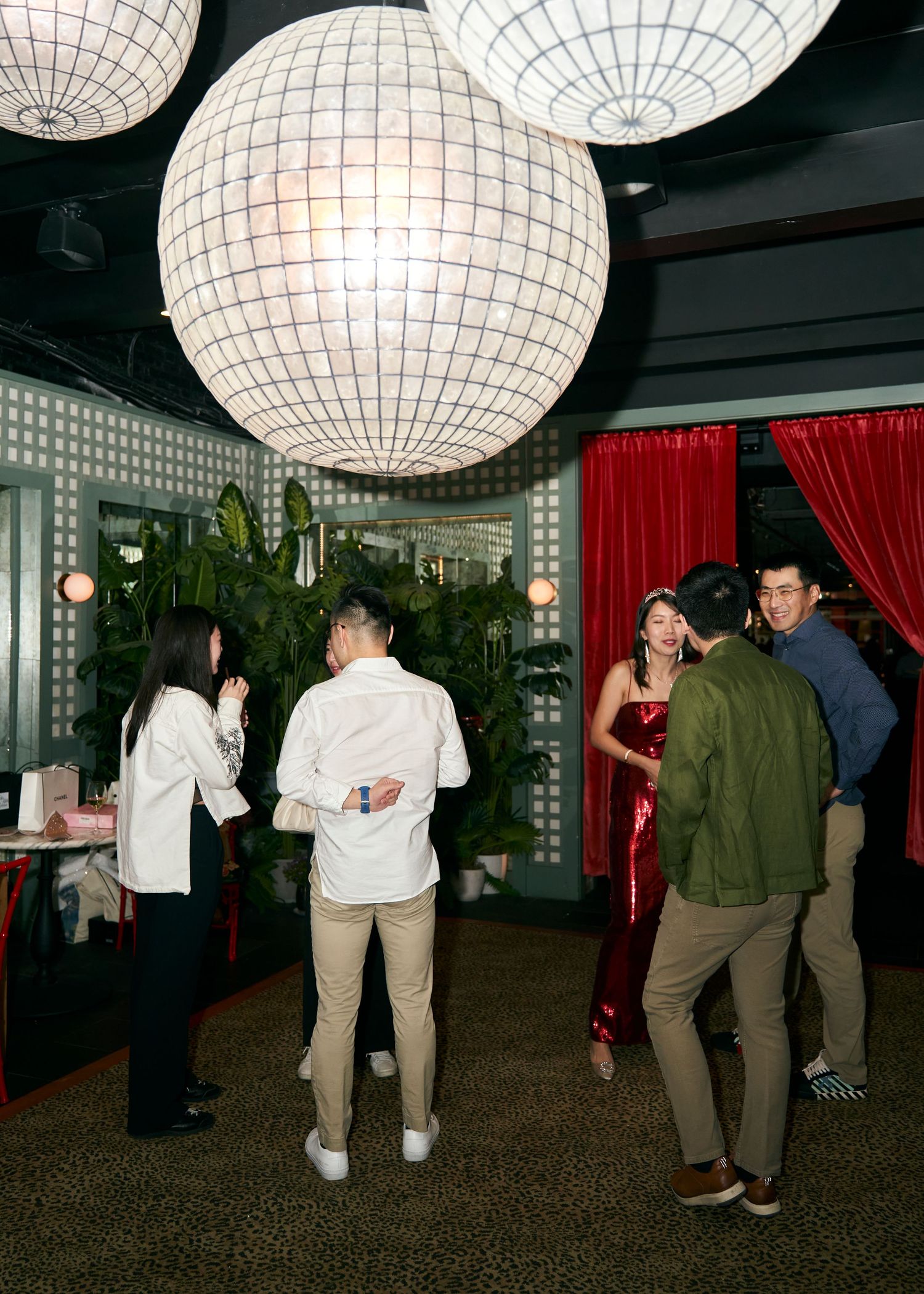 Guests mingle at a stylish birthday party venue with large globe pendant lights and red curtains.