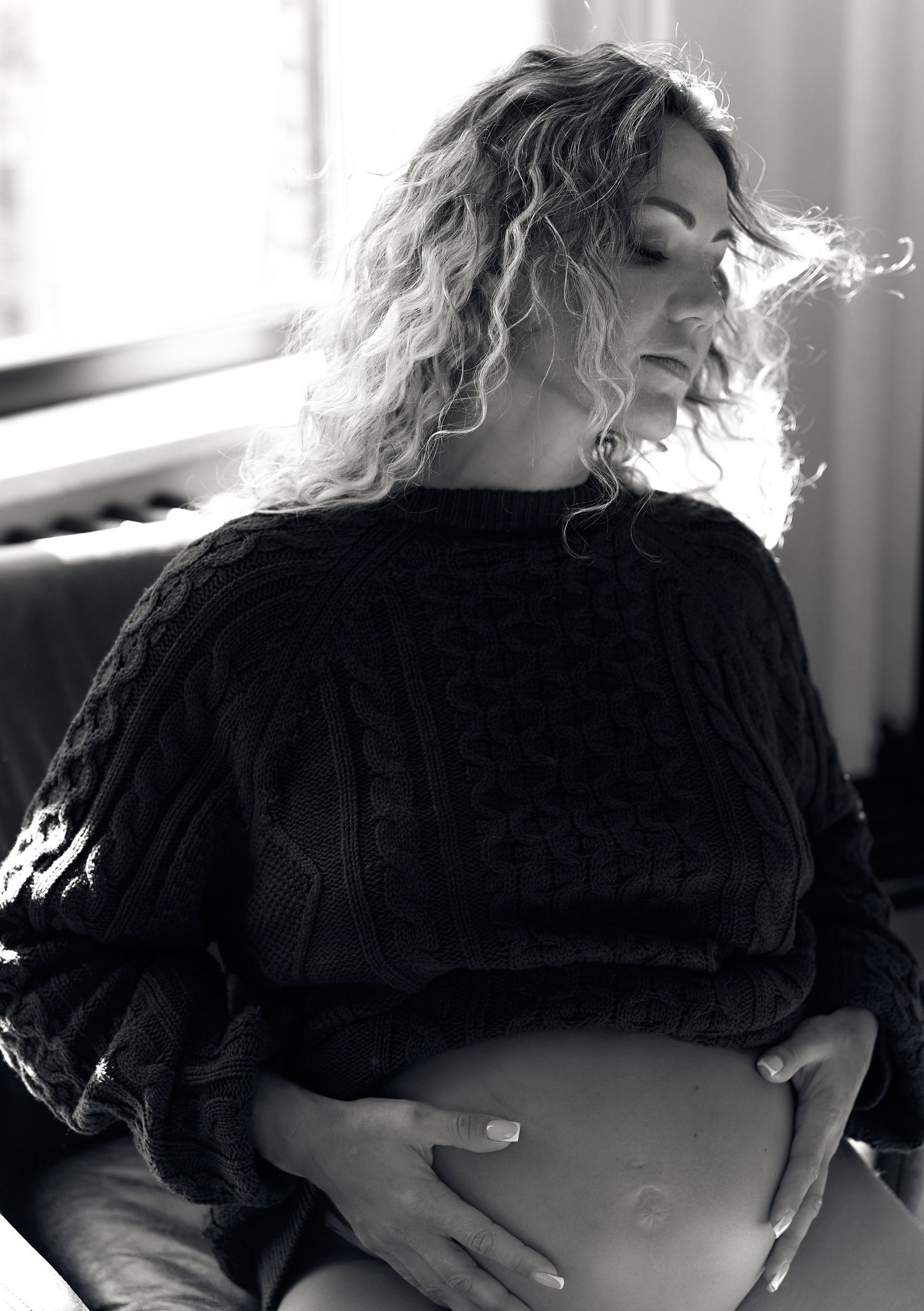 Curly-haired pregnant woman in black cable-knit sweater gently holds her bare bump in soft light.