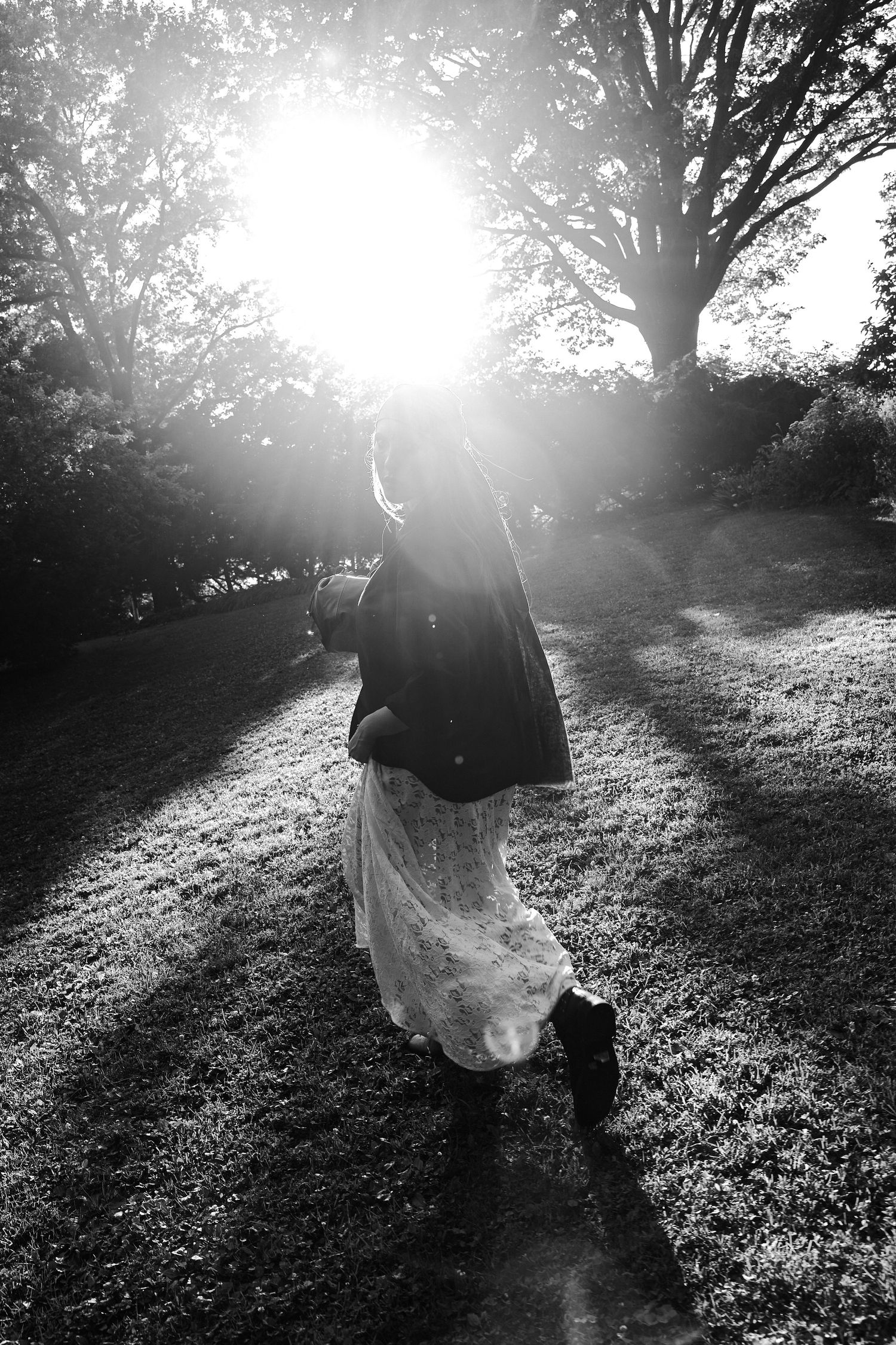 Woman in lace skirt and jacket walking through sunlit park with dramatic lens flare effect.