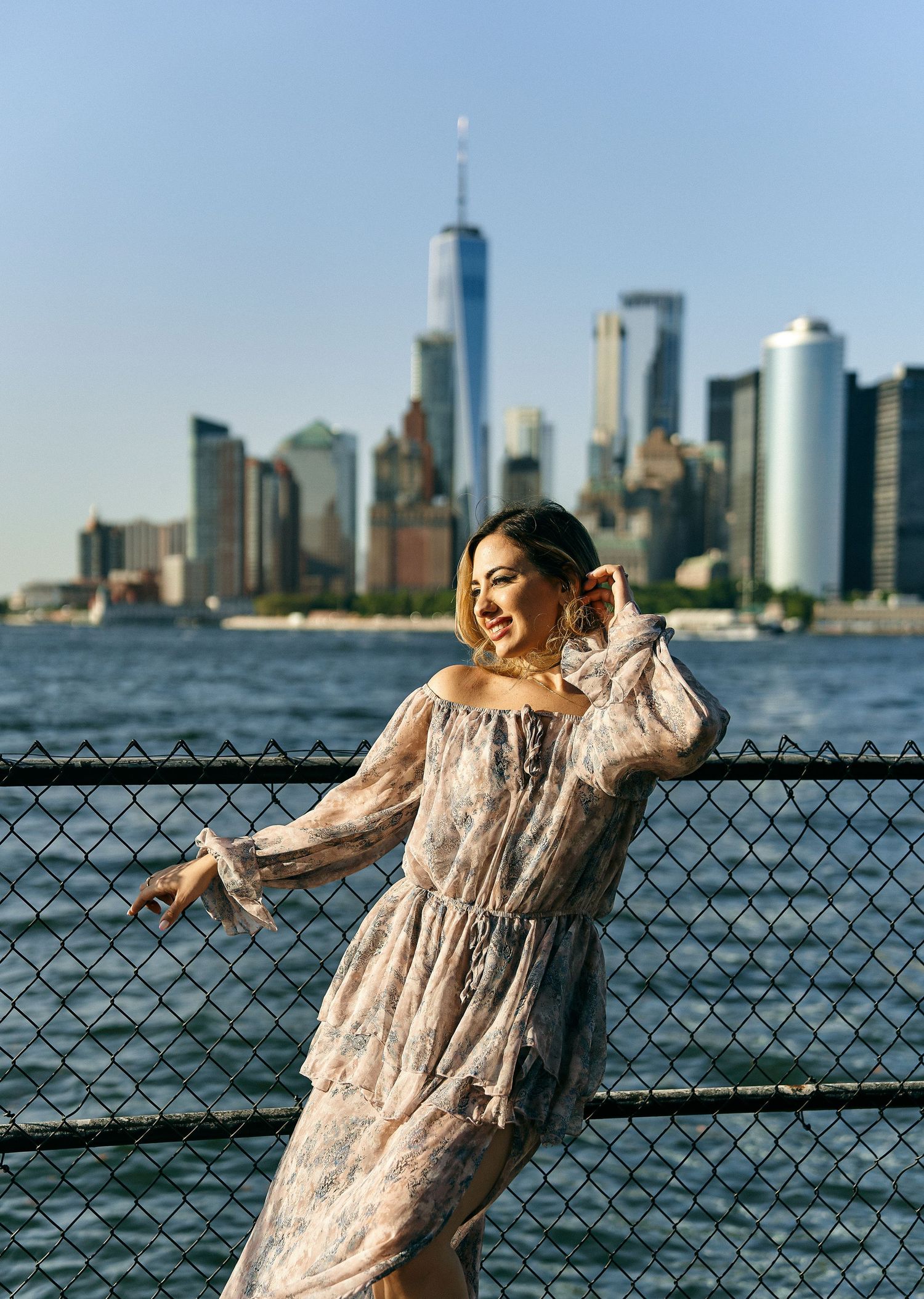 Woman in floral maxi dress posing by fence with NYC skyline backdrop.