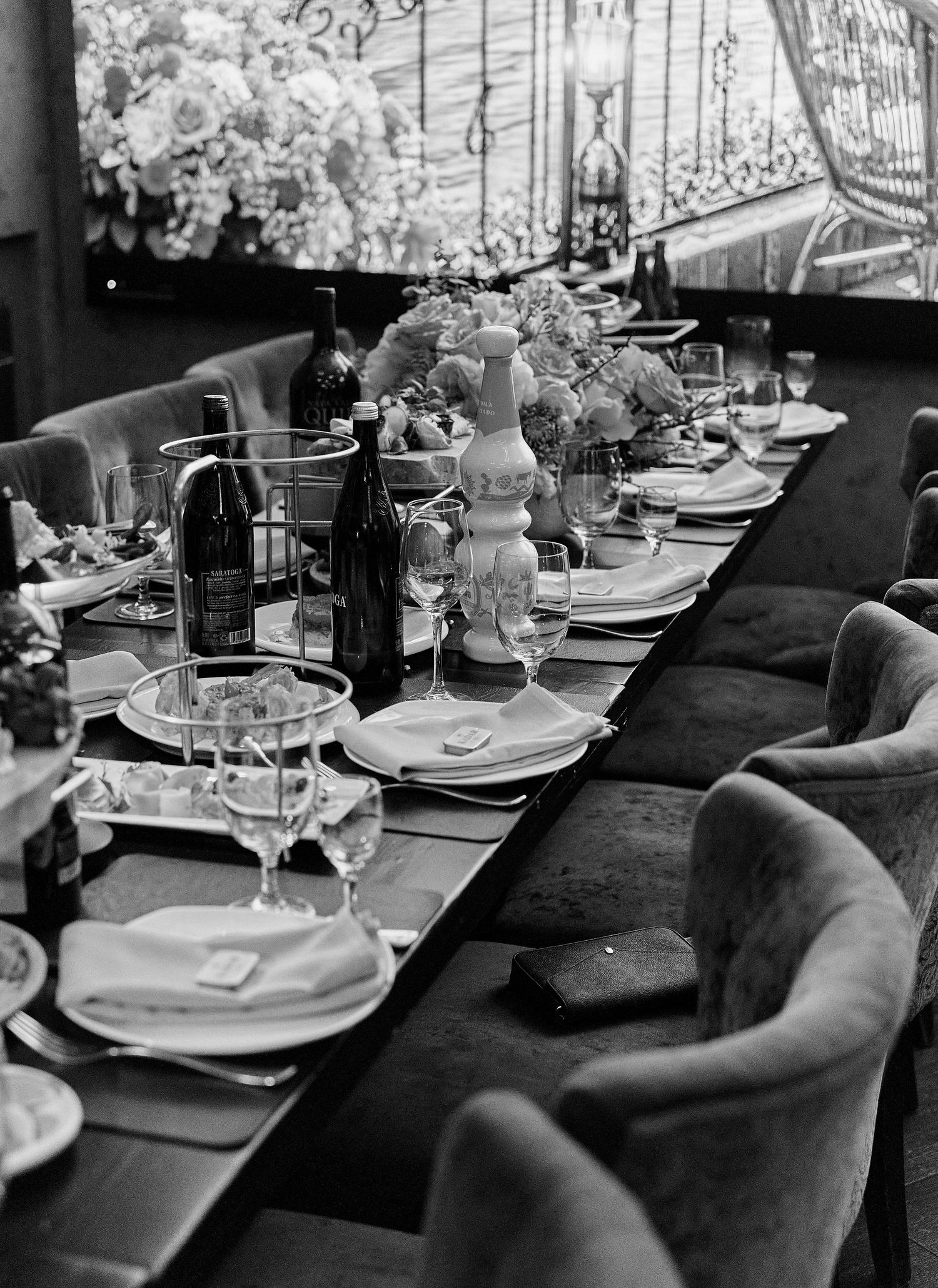 Black and white photo of a long elegantly set dining table with velvet chairs and floral centerpieces.