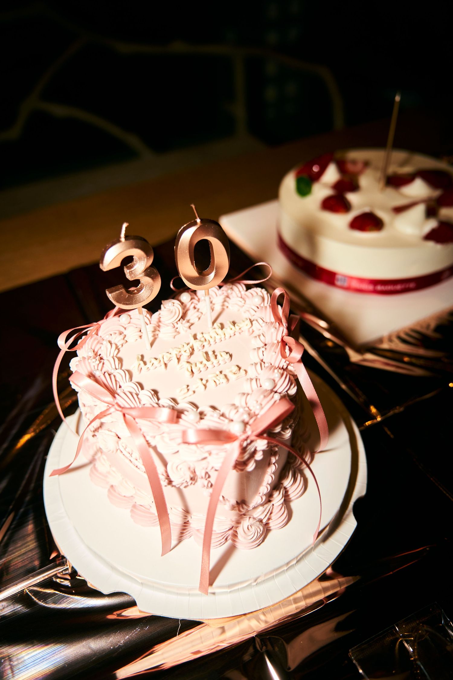 Heart-shaped 30th birthday cake with pink ribbons and candles beside a white cherry decorated cake.