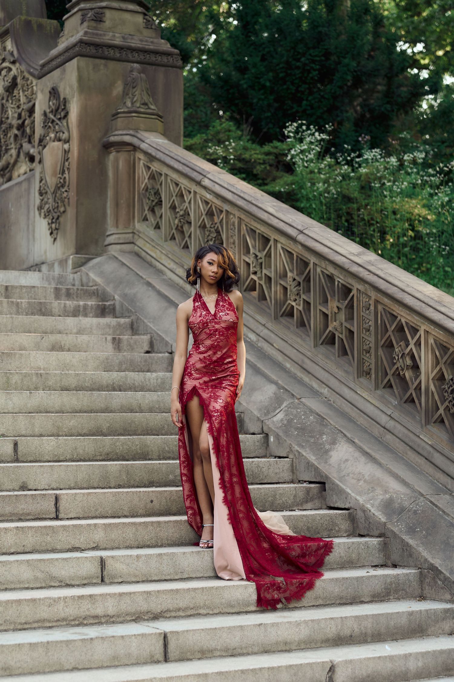 Woman in red lace gown with slit poses on ornate stone staircase outdoors.