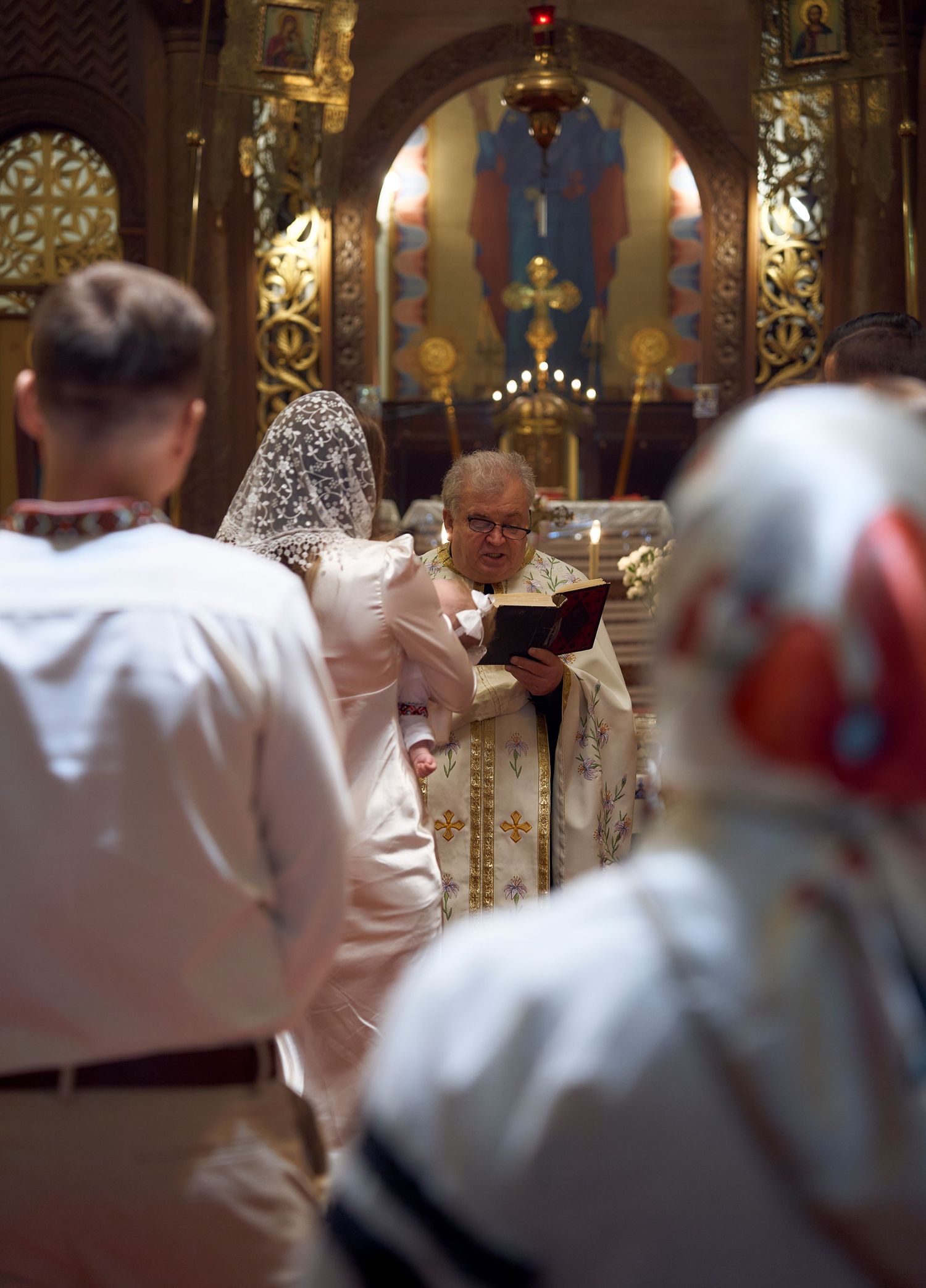 Orthodox priest in gold vestments reading from a book during a baptism ceremony in an ornate church.