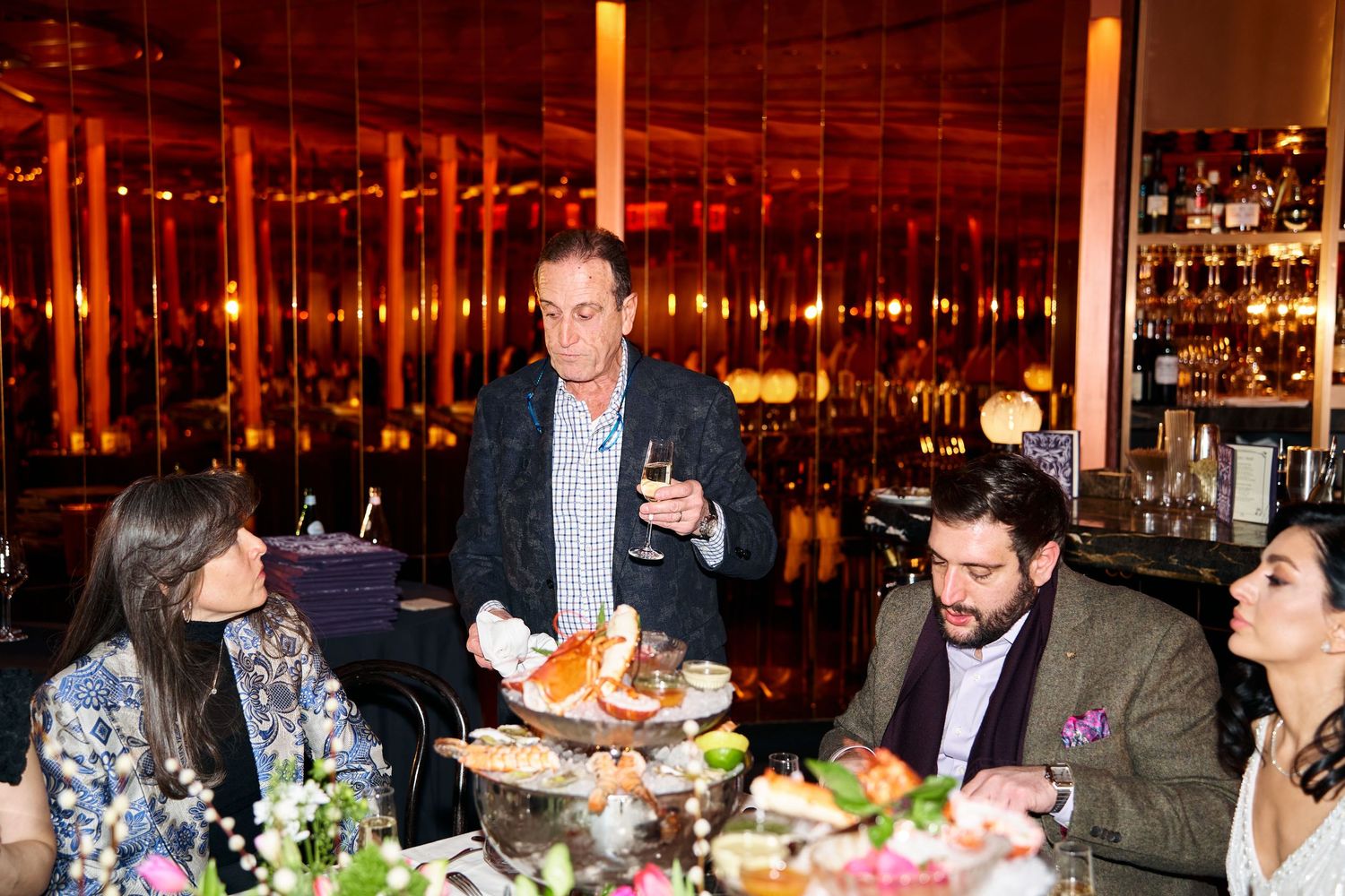 Man standing and speaking with a wine glass at an elegant restaurant dinner table with guests seated.