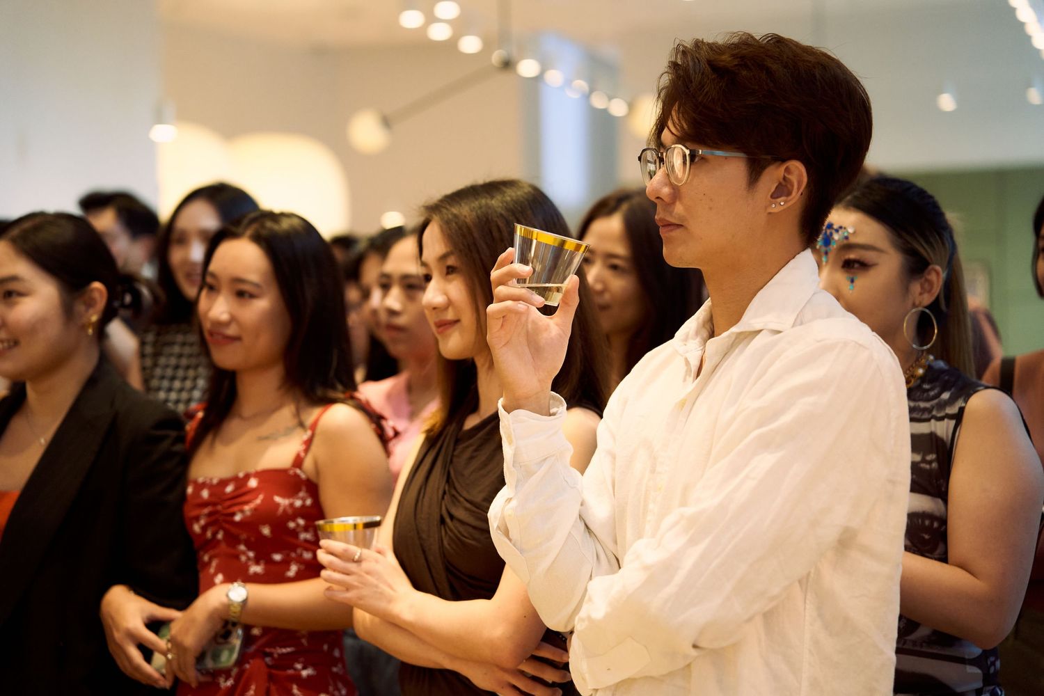 Man in white shirt holds drink at crowded art gallery social event.
