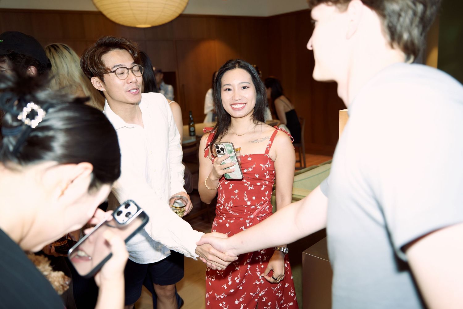 Young woman in red floral dress smiles while holding a drink and chatting at an indoor party.