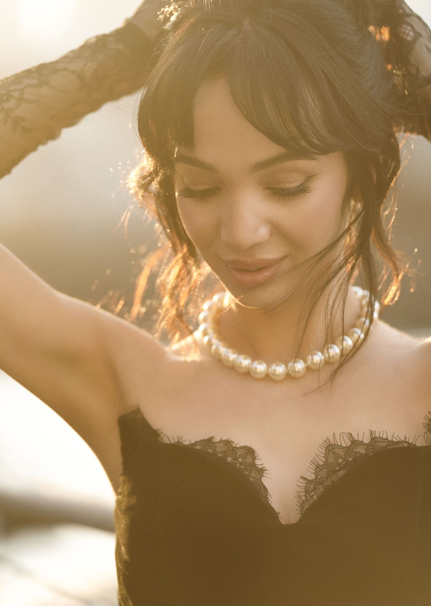Woman in black lace strapless gown wearing pearl necklace poses in golden sunlight.