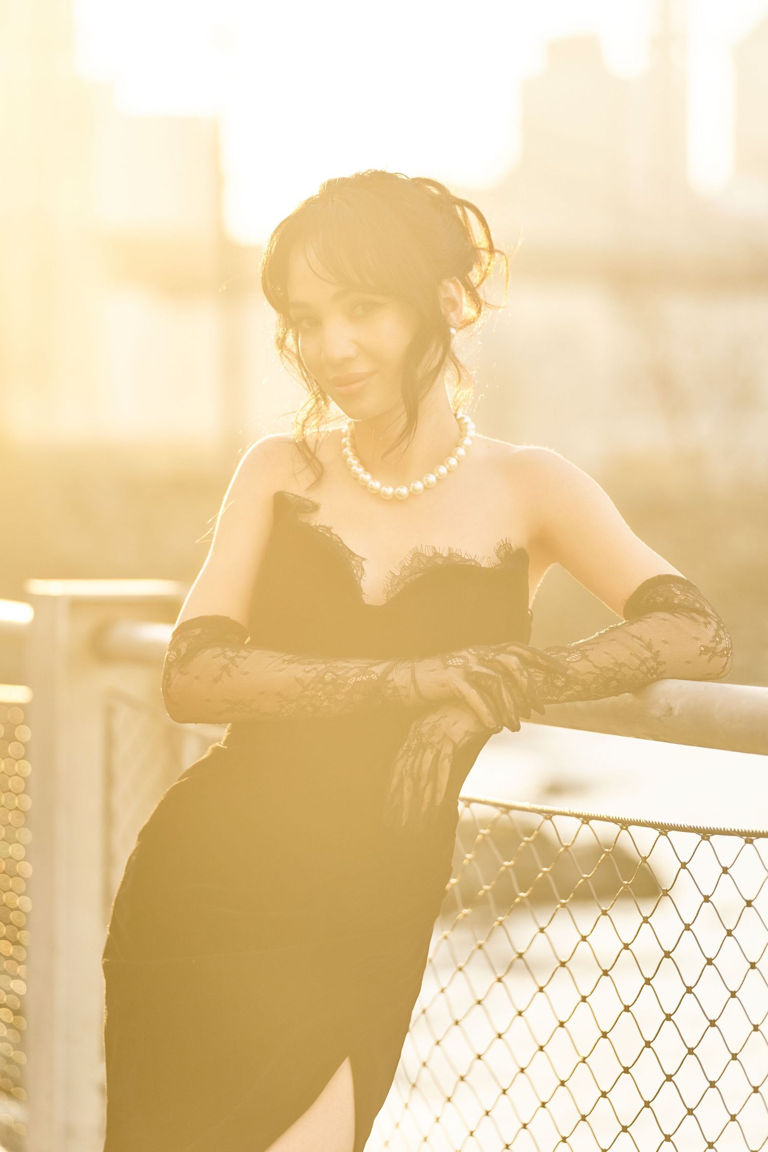 Elegant woman in black strapless gown with lace gloves leaning on railing at golden hour.