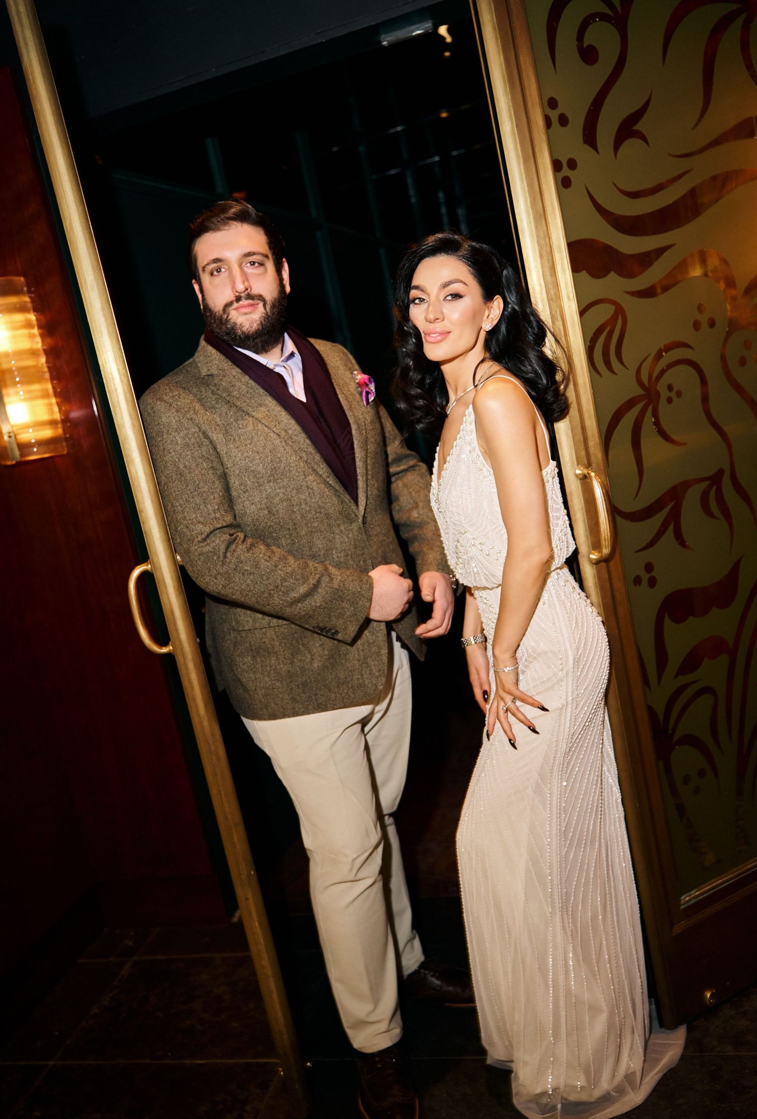 Stylish couple posing in a doorway, man in tweed blazer and woman in elegant beaded white dress.