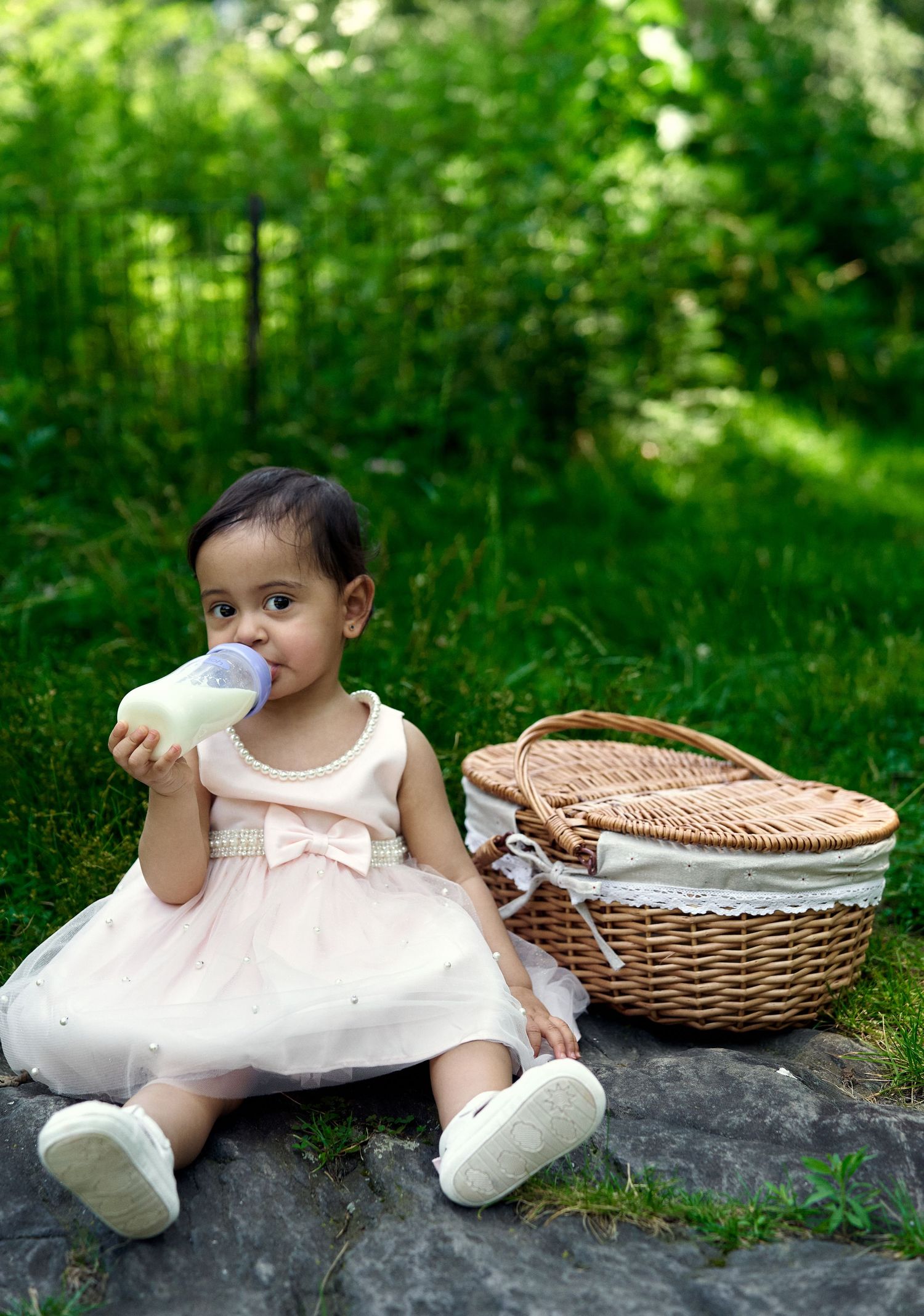 Toddler in pink dress drinking from bottle beside wicker basket in park.