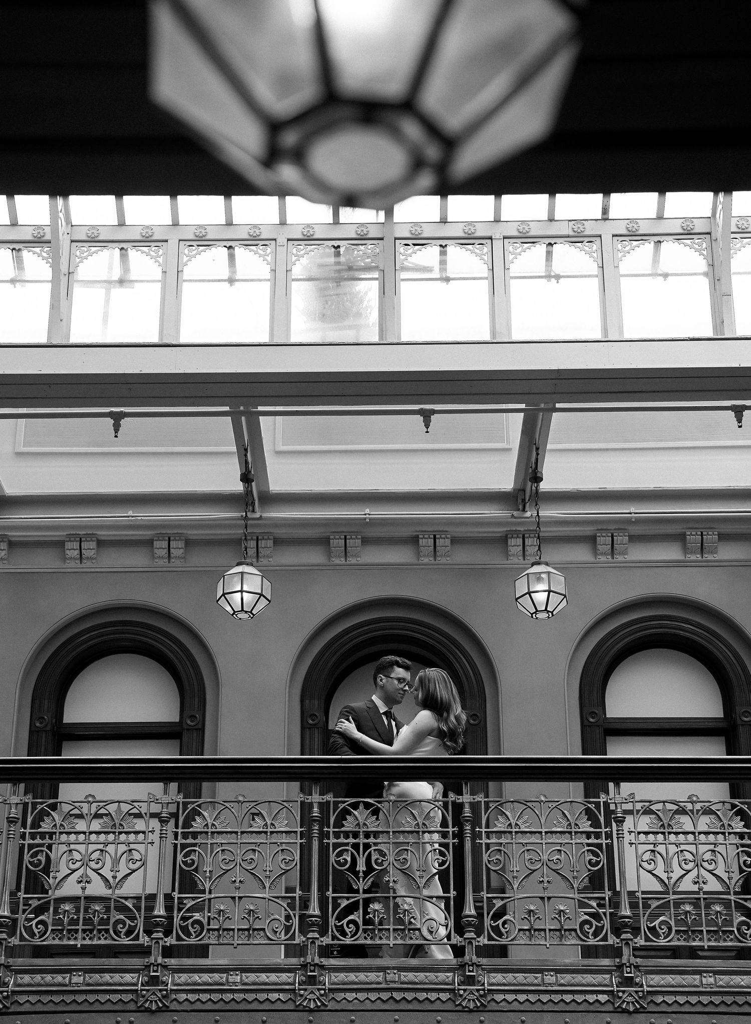 Black and white photo of couple kissing on ornate balcony inside elegant historic building.