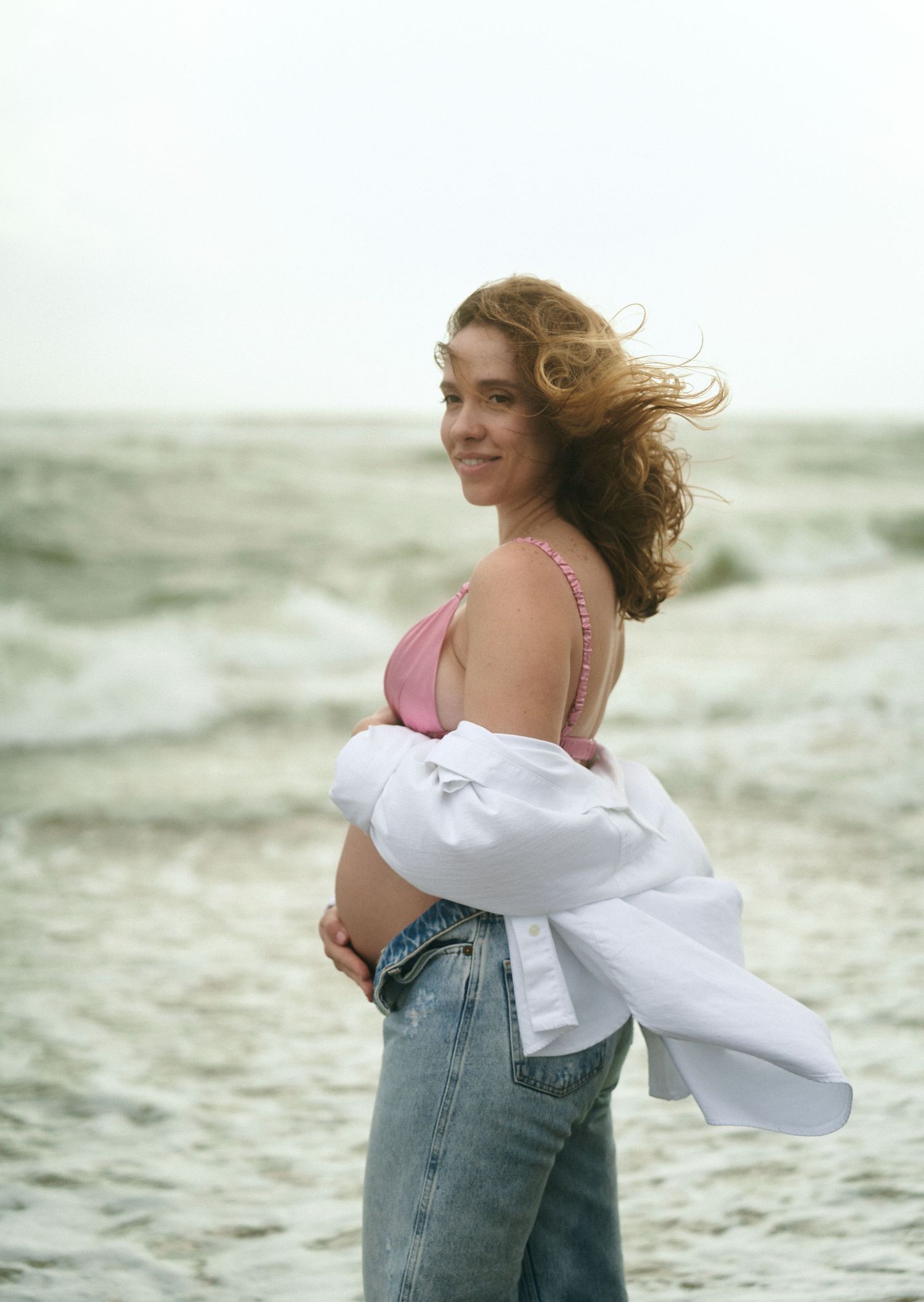 Smiling pregnant woman on beach wearing pink top and open white shirt with jeans.