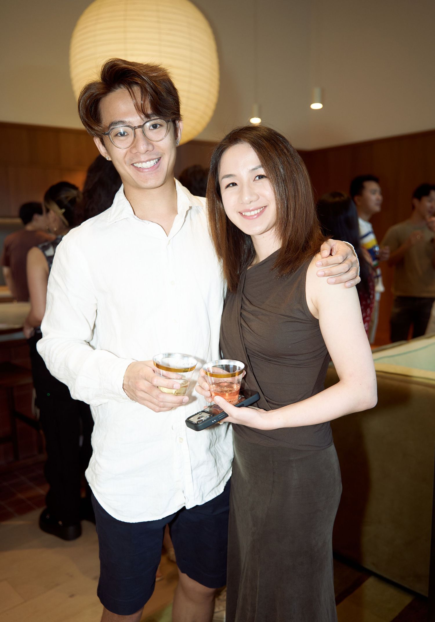 Smiling couple poses together holding cocktail glasses at a casual indoor social gathering.