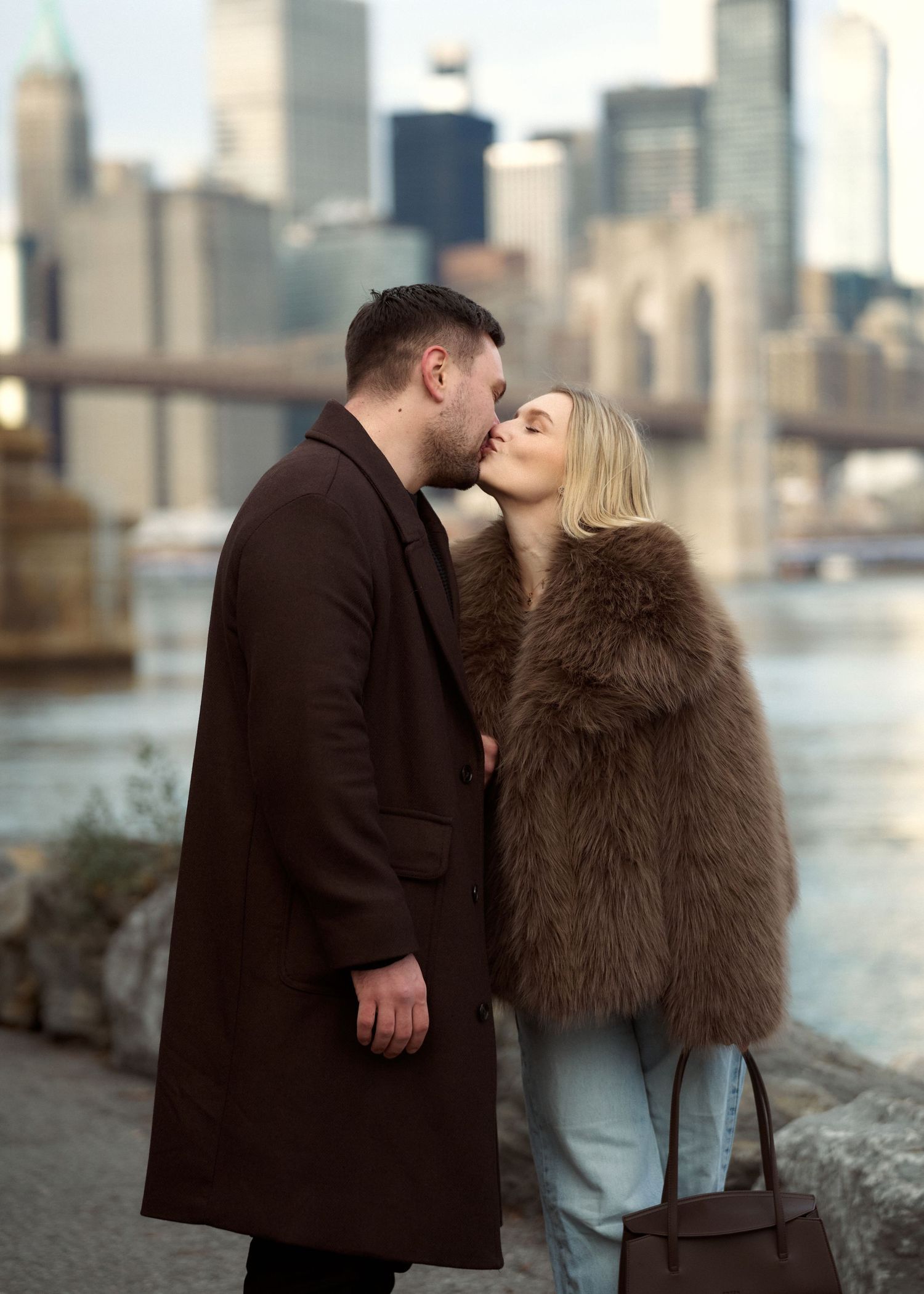 Couple kissing near the Brooklyn Bridge waterfront, woman in fur coat and man in brown overcoat.