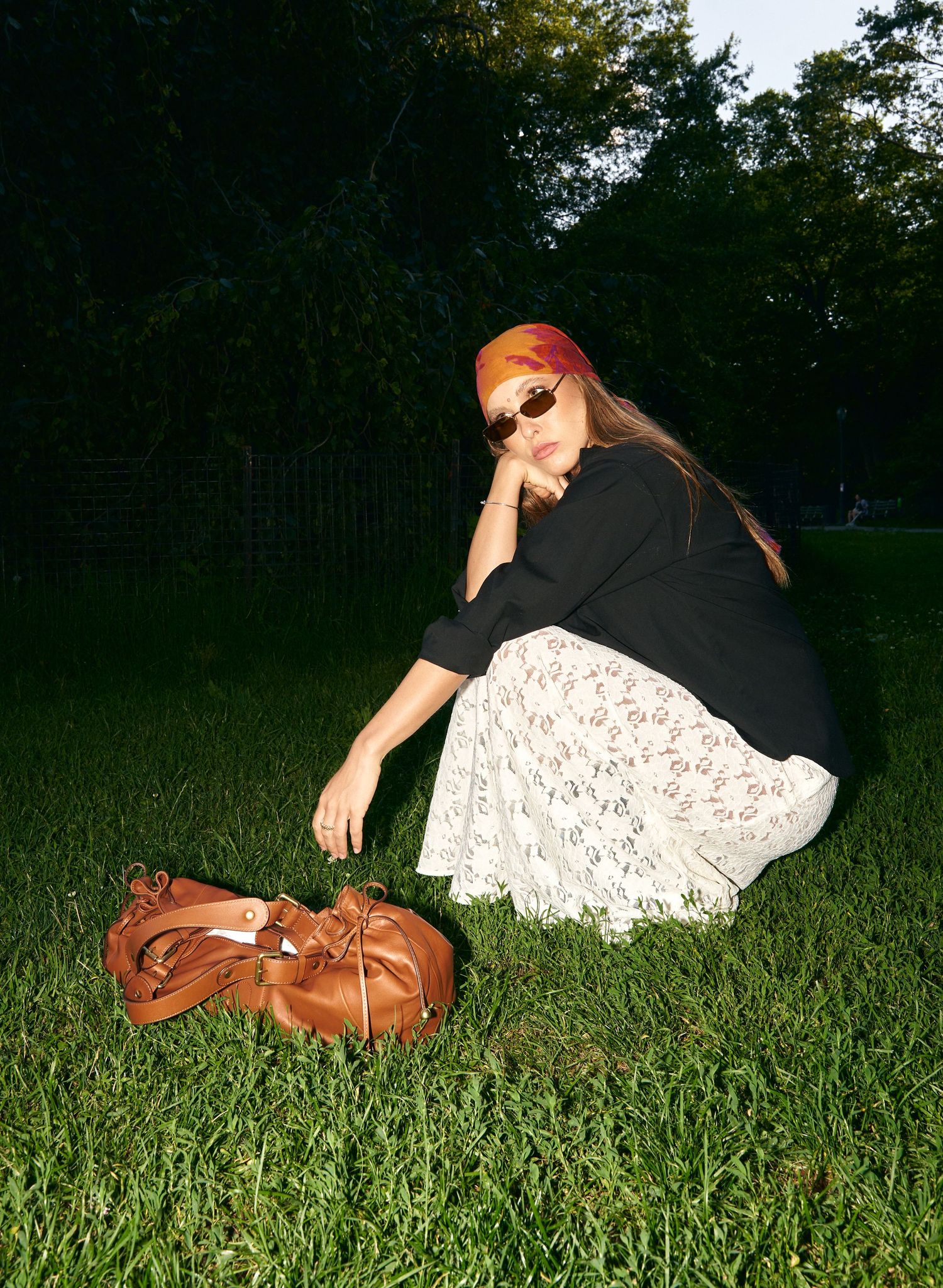 Stylish woman in white lace skirt and black top reaching for brown leather bucket bag on grass.