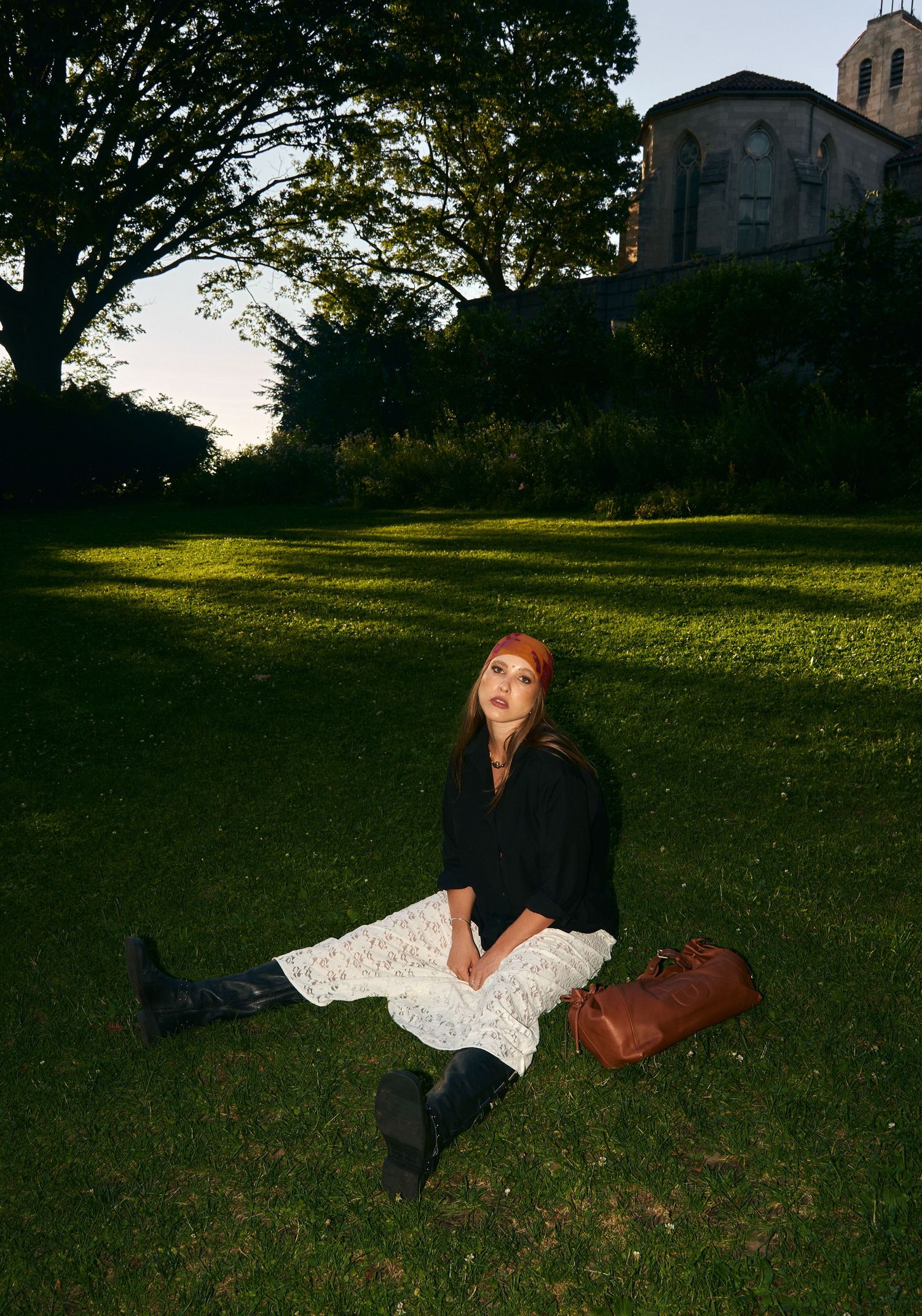 Woman in white lace skirt sitting on grass near brown leather bag with historic building behind.