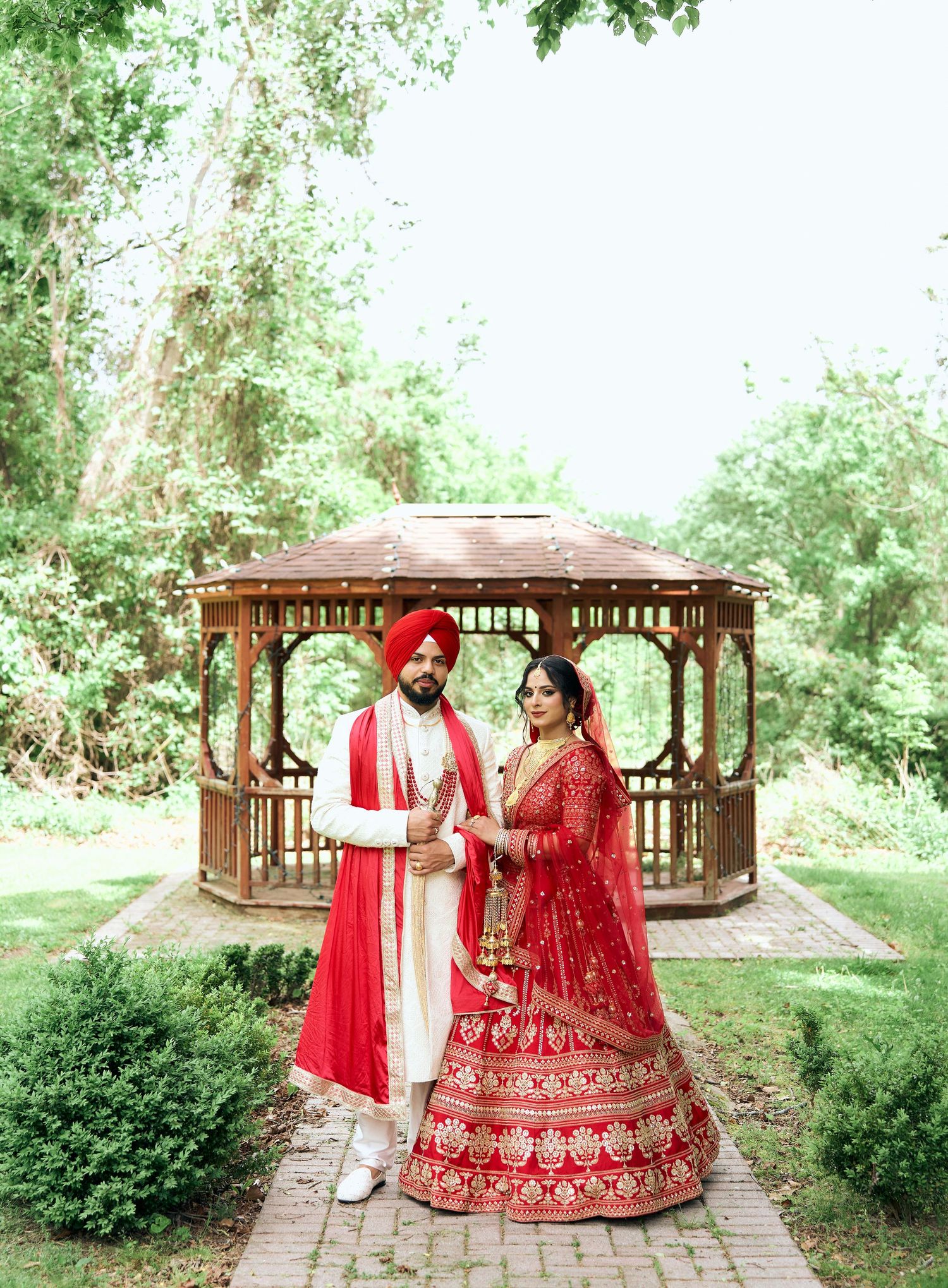 Sikh bride and groom in red traditional wedding outfits posing by wooden gazebo in garden.