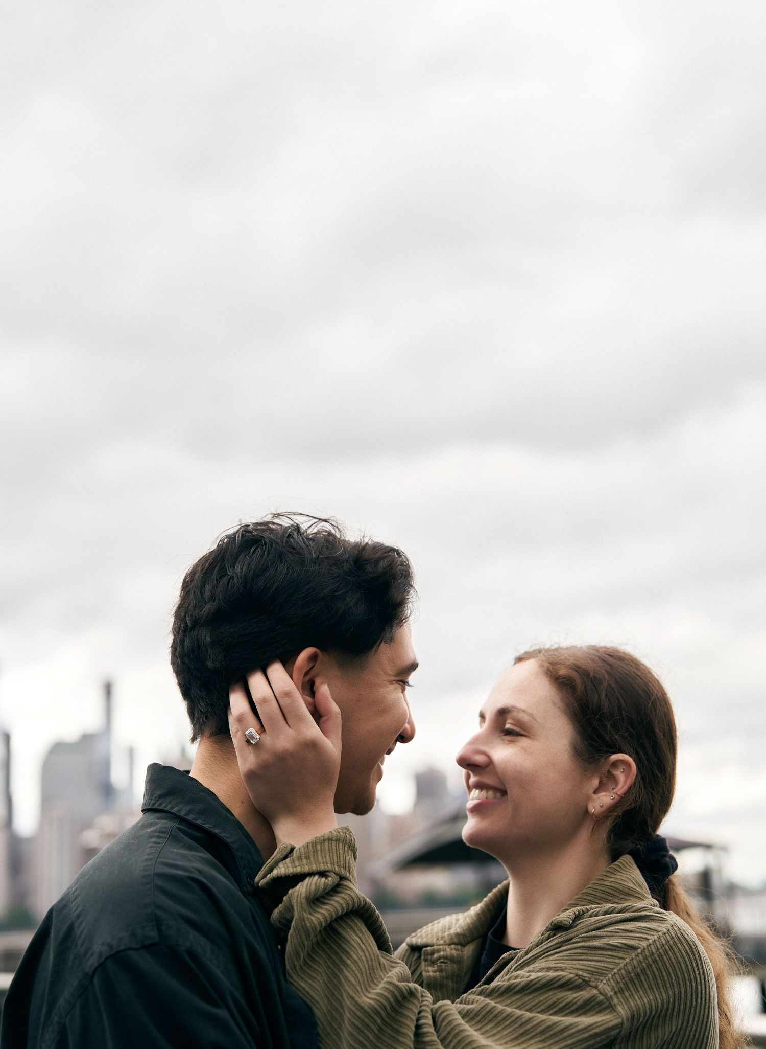 A couple shares a tender moment with a city skyline backdrop on an overcast day.