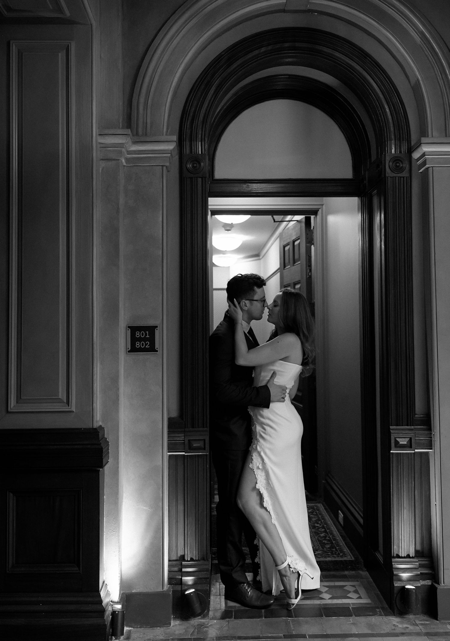 Black and white photo of couple embracing in arched doorway of elegant historic building interior.