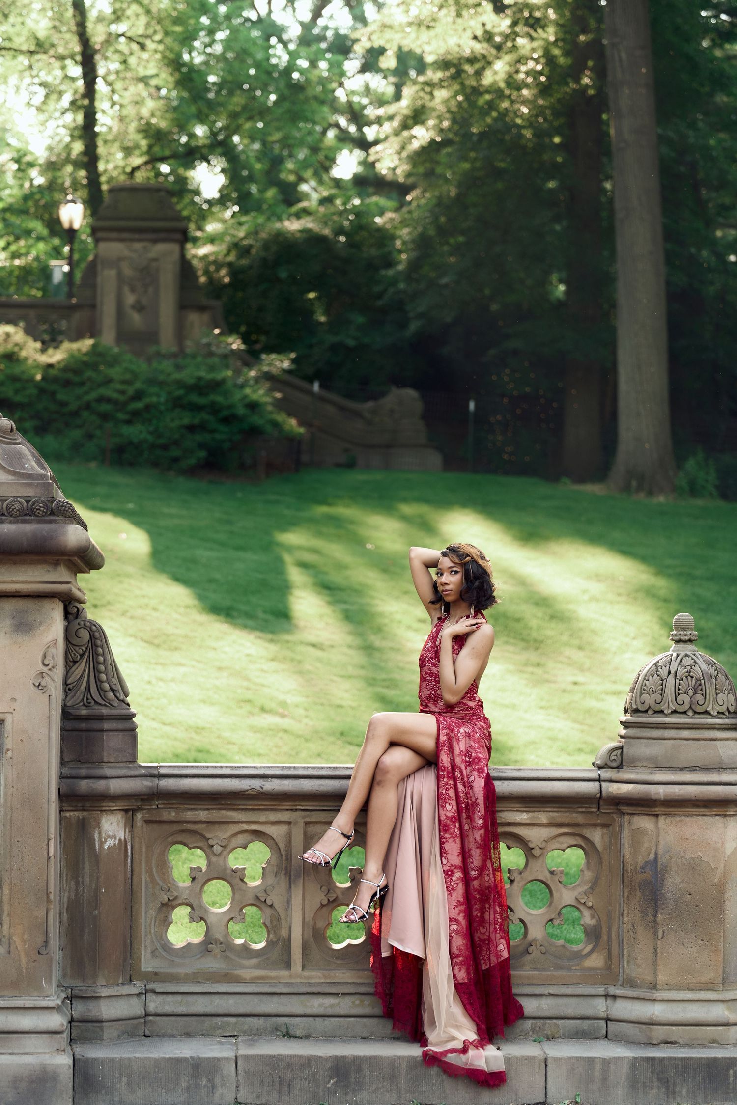 Woman in red lace dress sits on Bethesda Terrace stone railing surrounded by lush Central Park greenery.