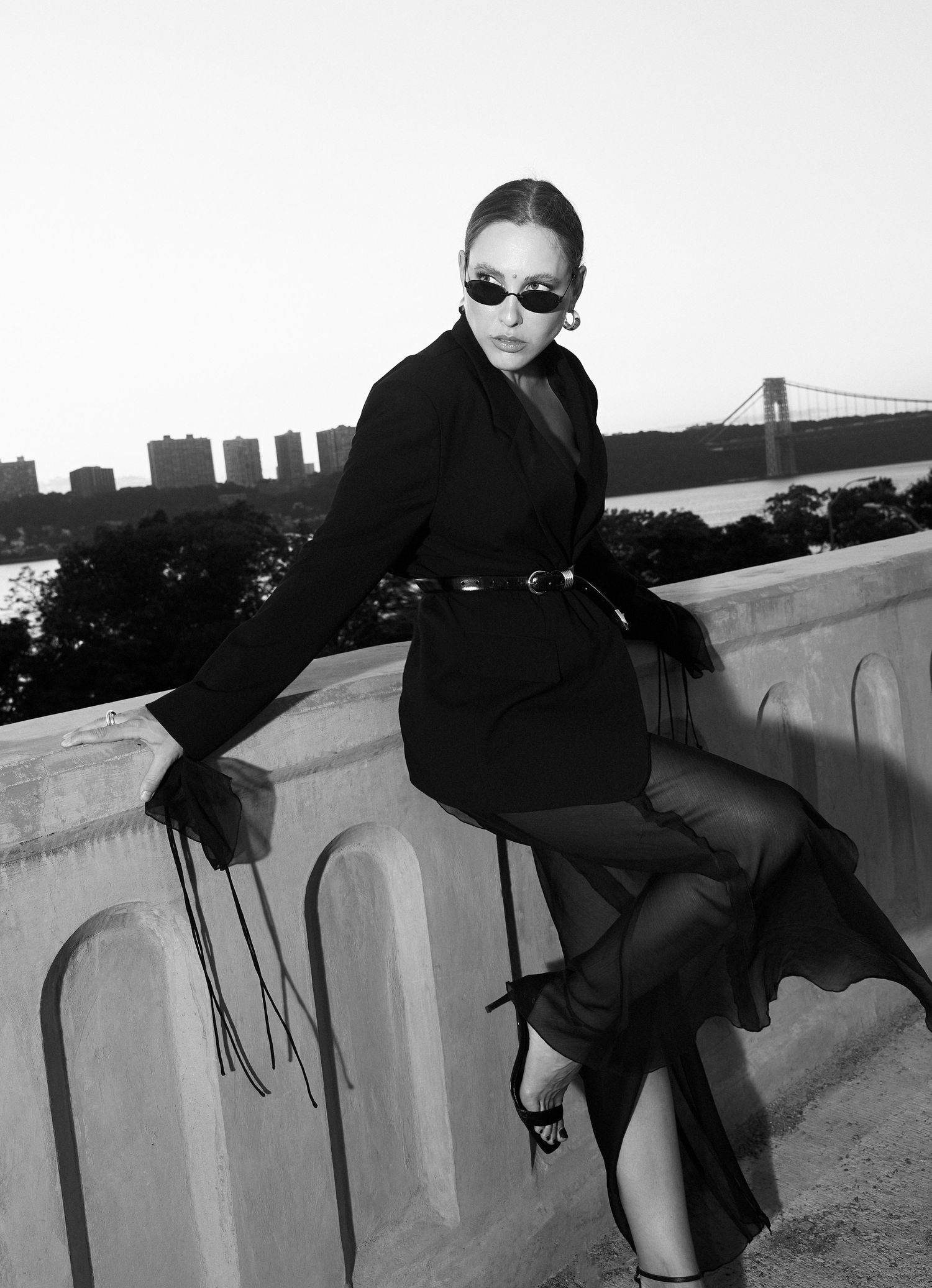 Black and white fashion photo of woman in belted black coat and sheer skirt on bridge wall.