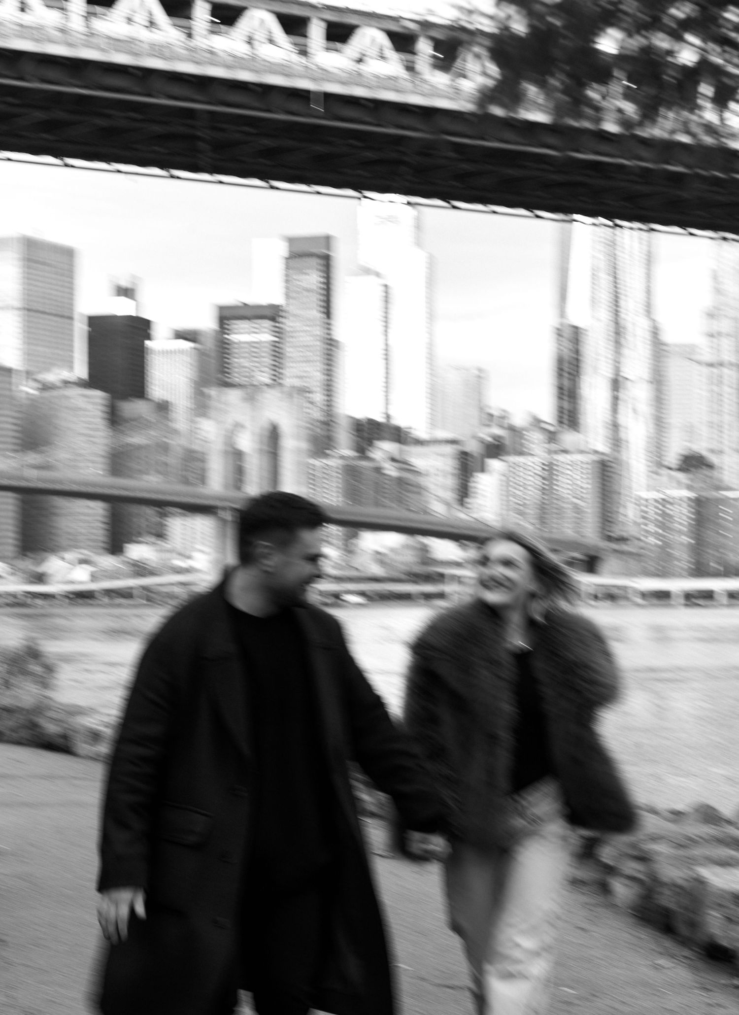 Black and white blurred motion photo of a couple holding hands and laughing under a bridge.