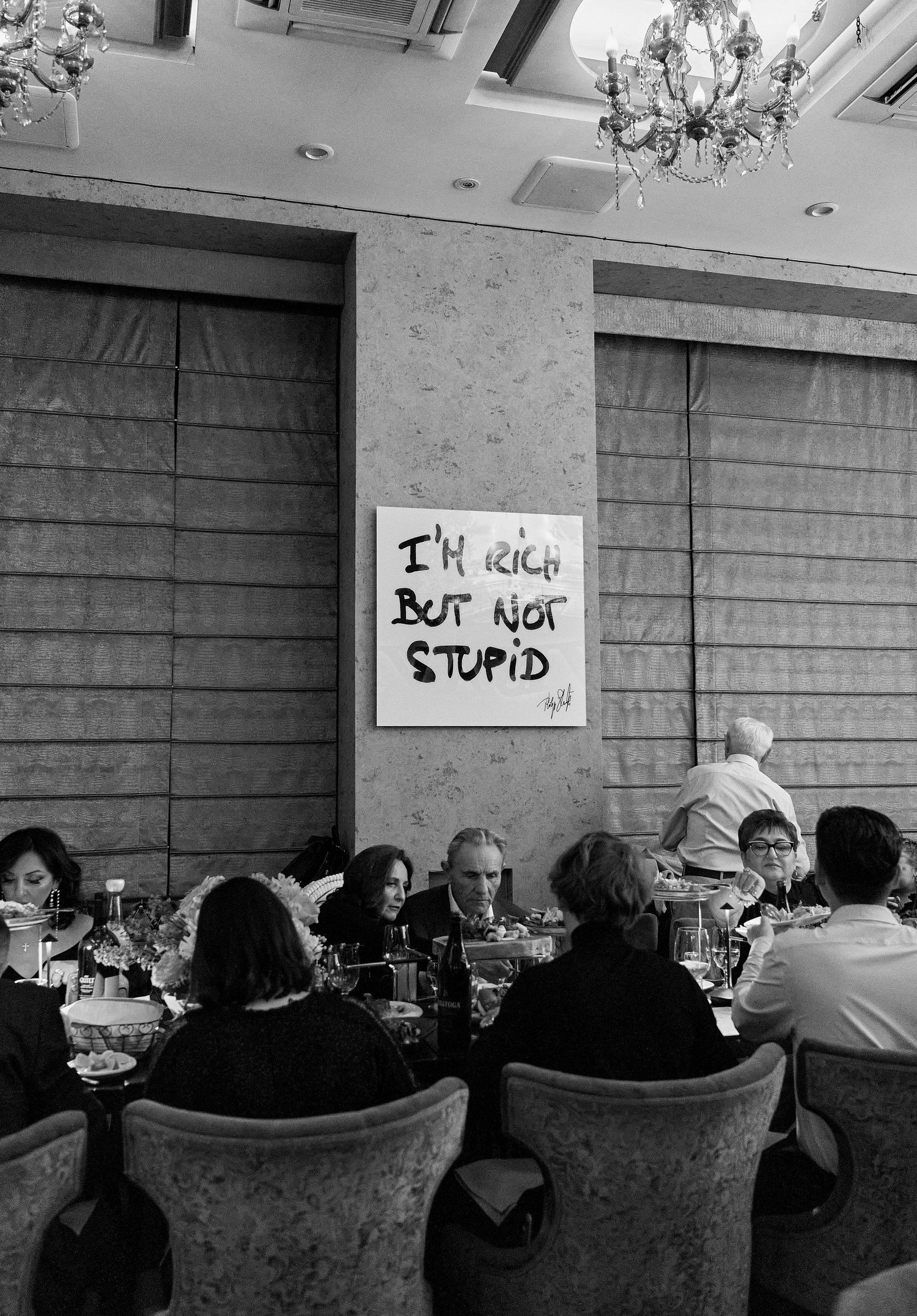 Black and white photo of diners at elegant restaurant beneath bold art reading I'm Rich But Not Stupid.