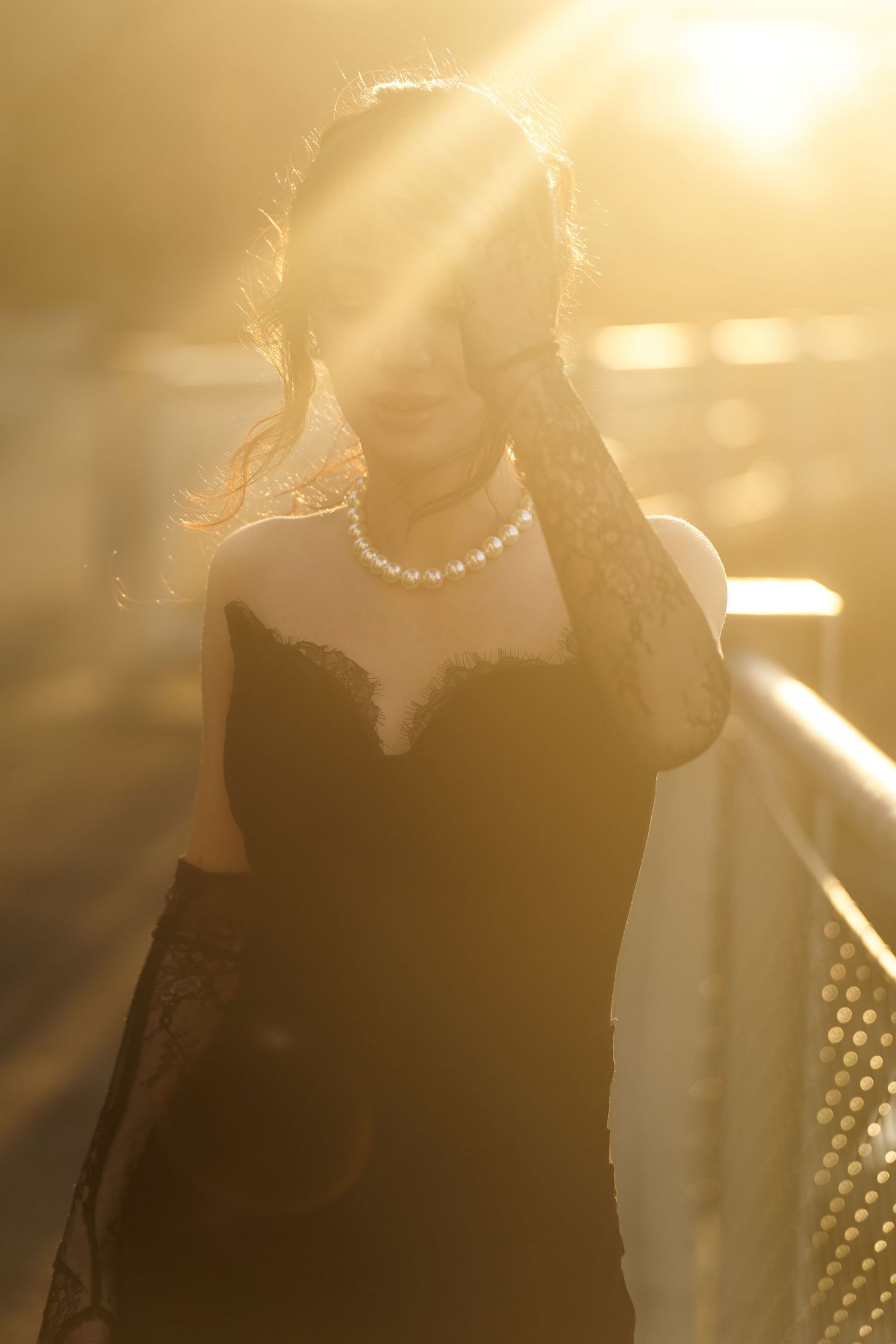 Woman in black lace dress and pearl necklace posing in golden sunset light on bridge.