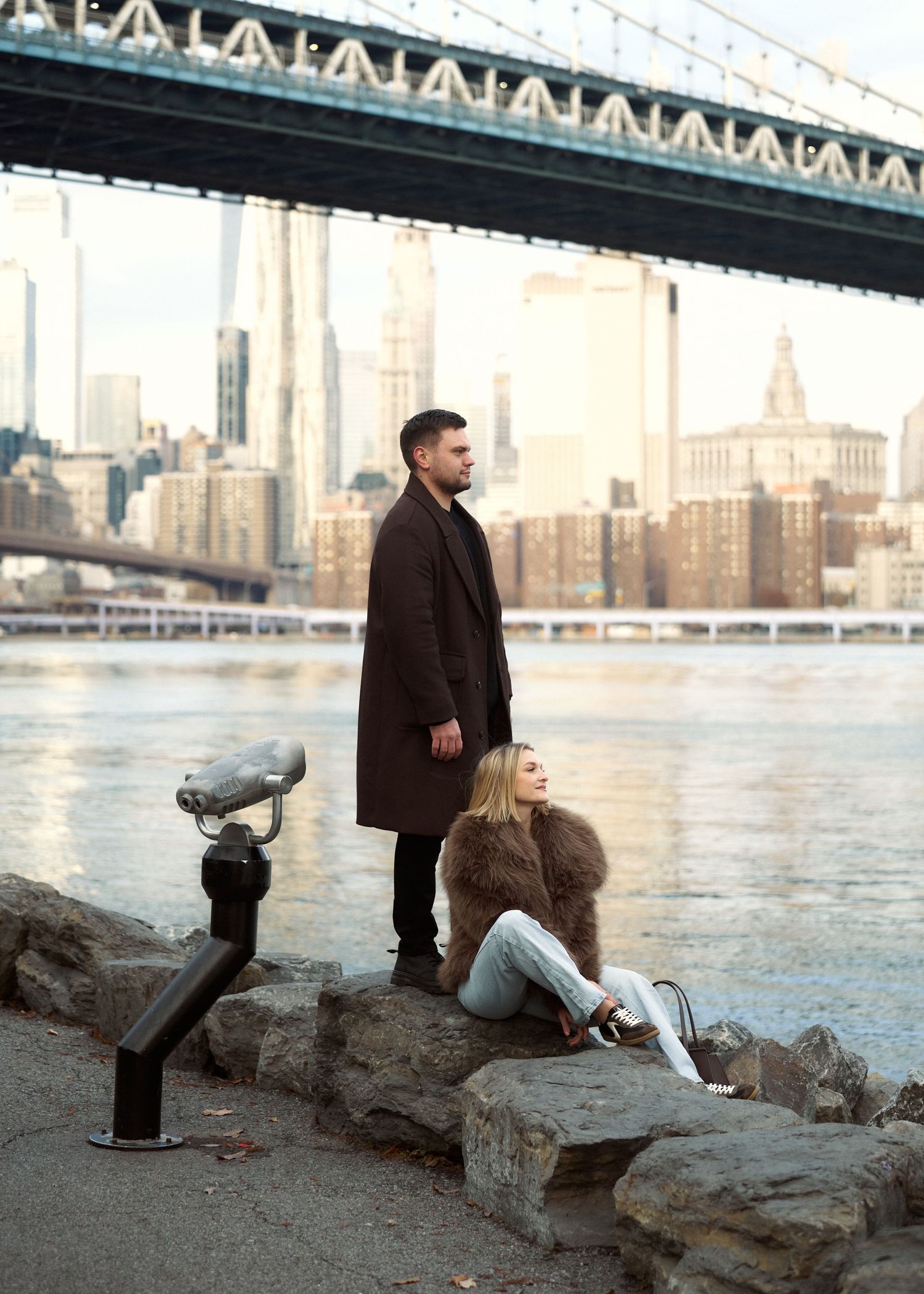 Couple posing by the Manhattan Bridge waterfront, man standing and woman sitting on rocks.