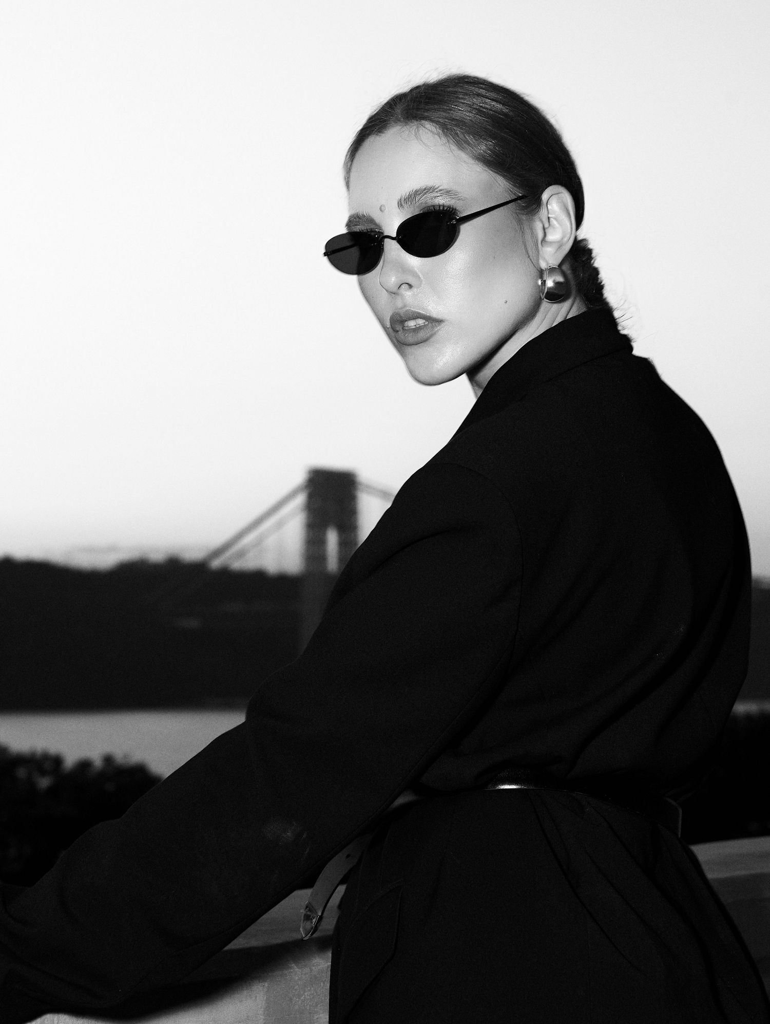 Black and white portrait of woman in sunglasses with bridge skyline in background at dusk.