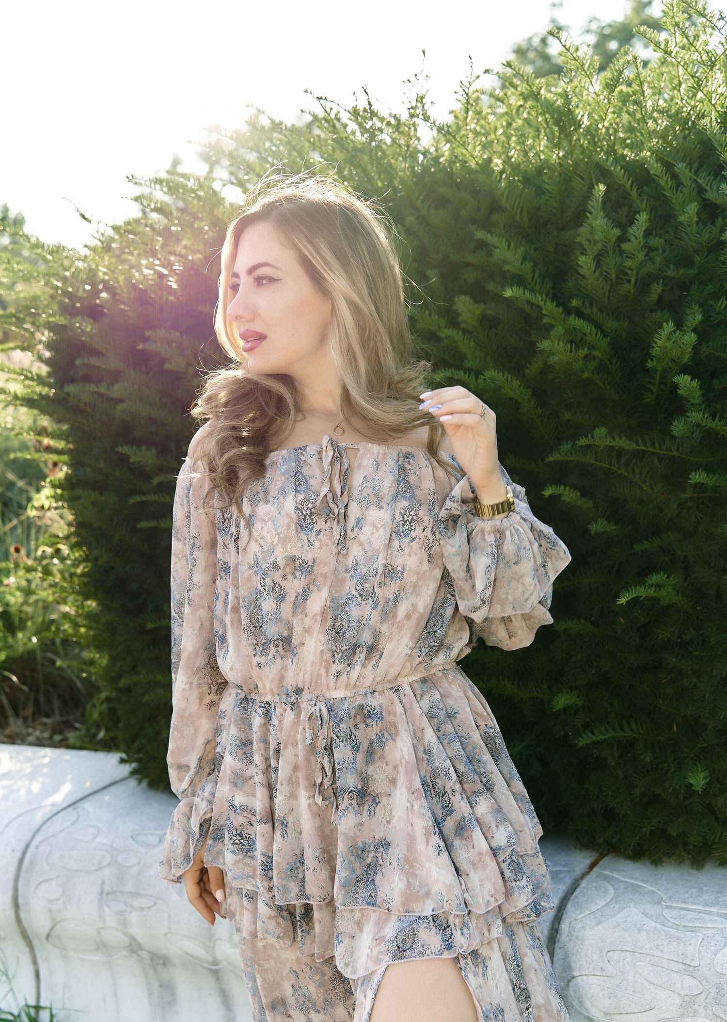 A blonde woman in a floral off-shoulder dress poses in a sunlit garden.
