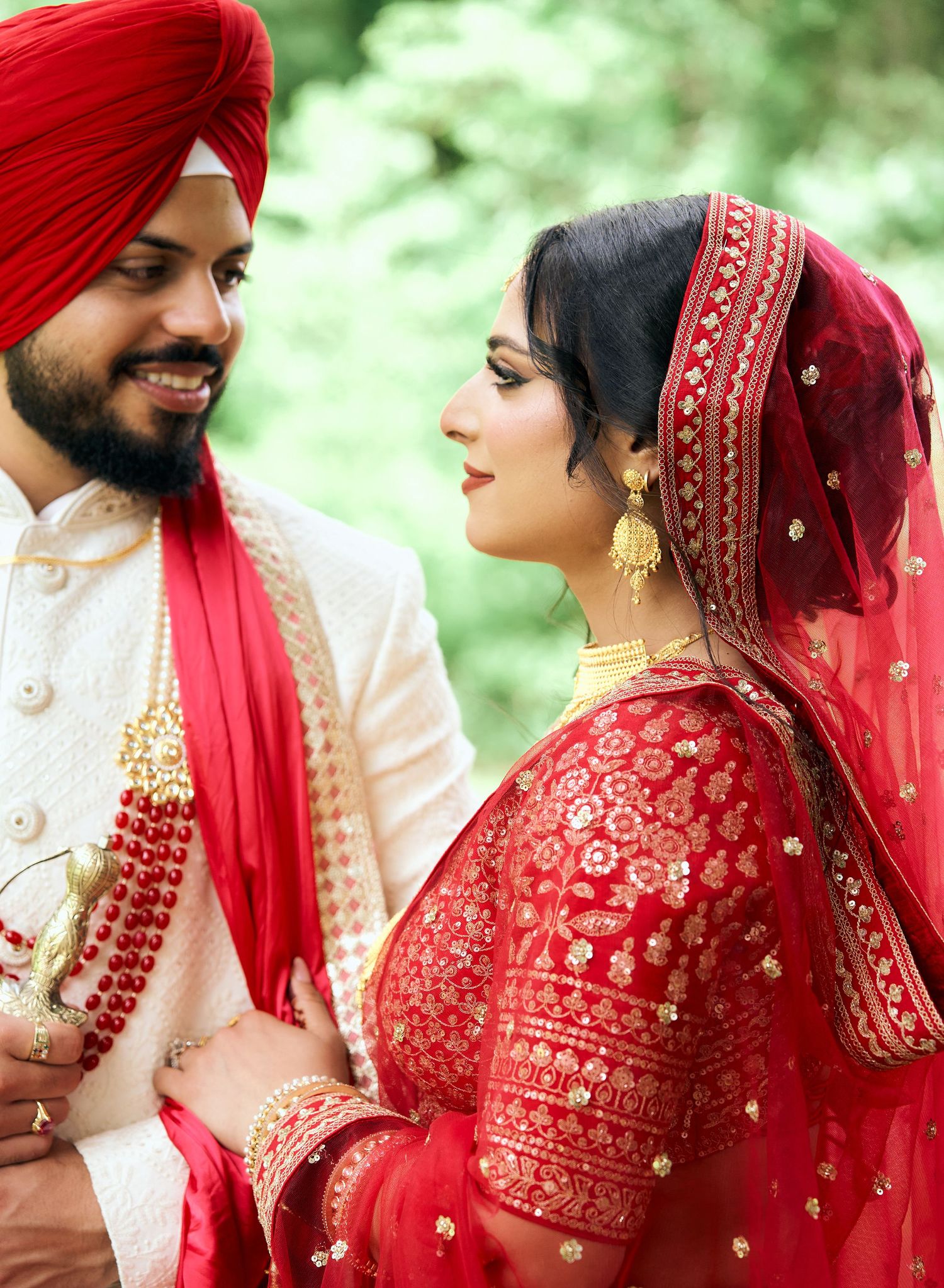 Sikh bride and groom in traditional red and gold wedding attire with turban and dupatta outdoors.