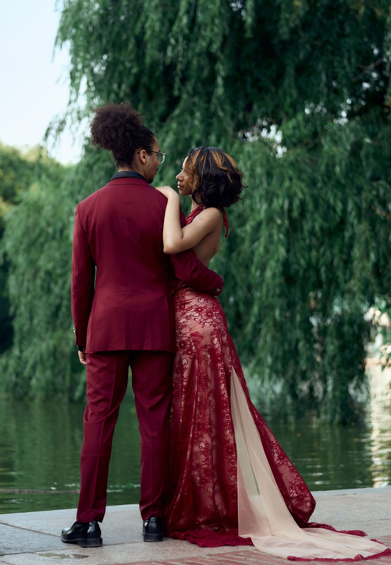 Couple in matching burgundy formal wear embrace by a serene lake with willow trees.