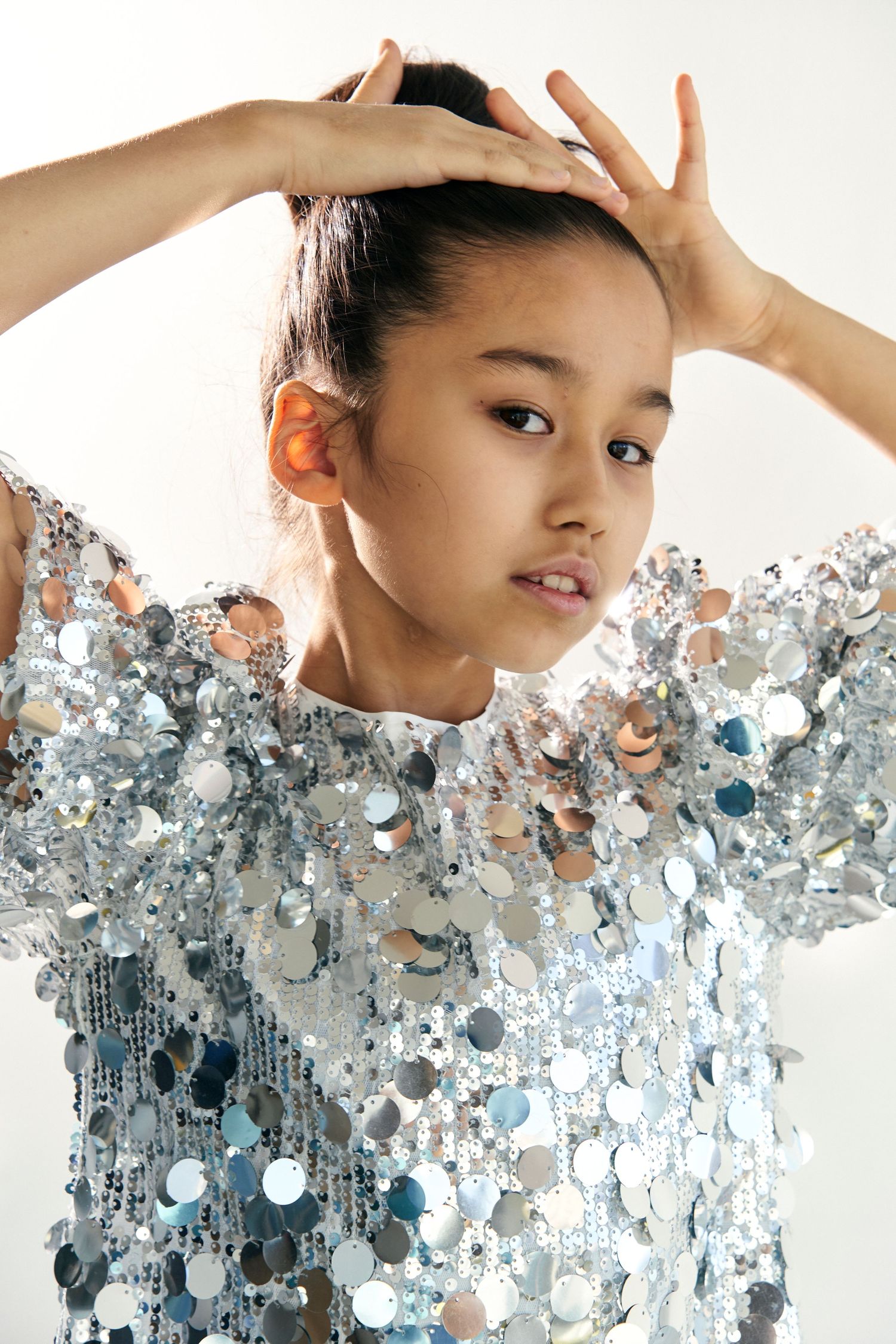 Young girl in silver sequin top poses with hands on head against white background.