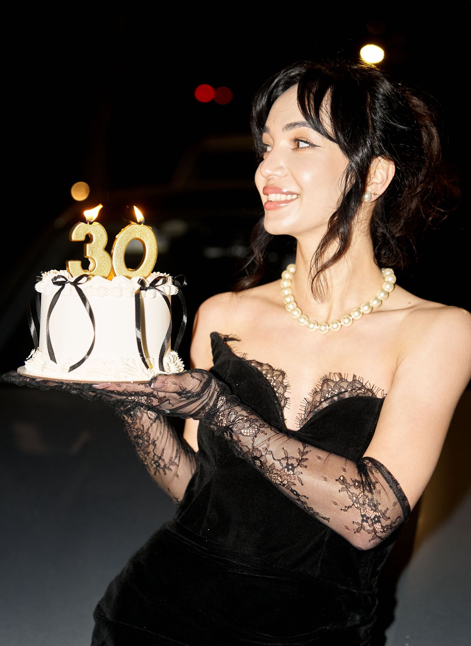 Smiling woman in black strapless gown holding a 30th birthday cake with gold candles at night.