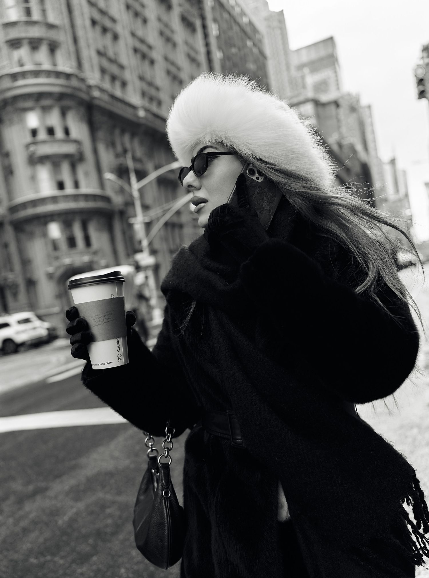 Black and white photo of a blonde woman in a fur headband and black coat holding a coffee cup.