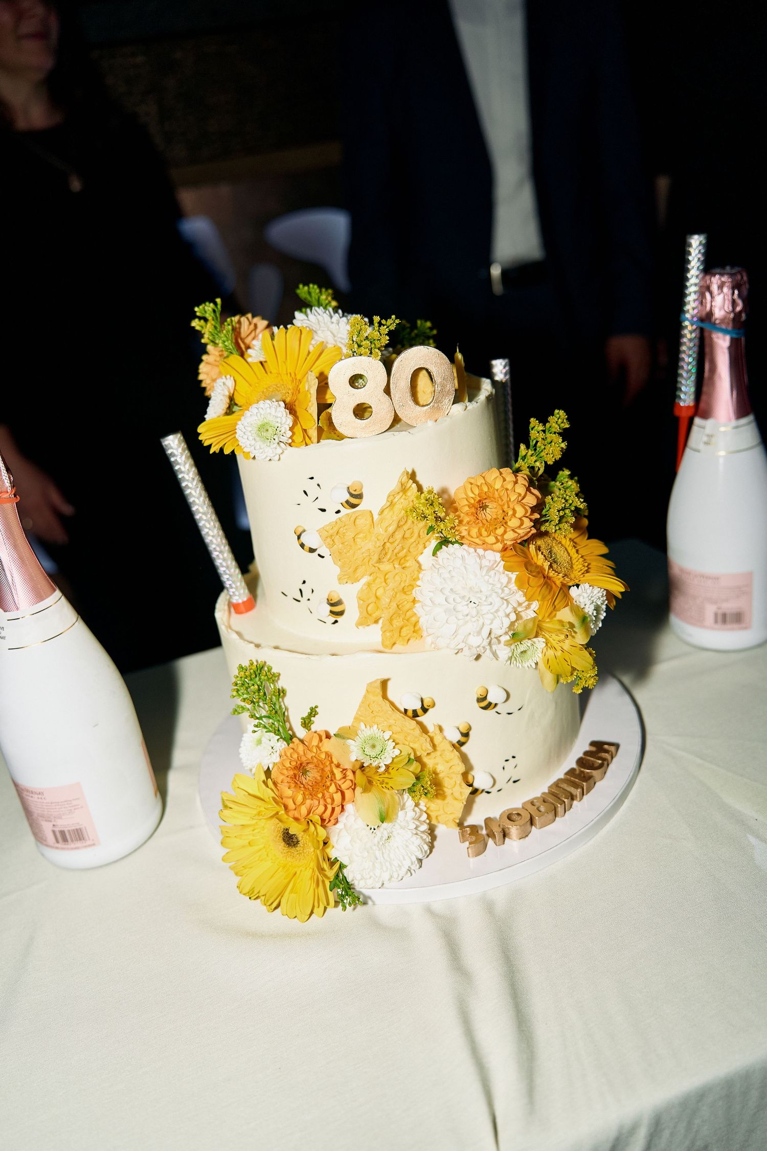 Two-tier white 80th birthday cake decorated with yellow and white flowers and gold number topper.