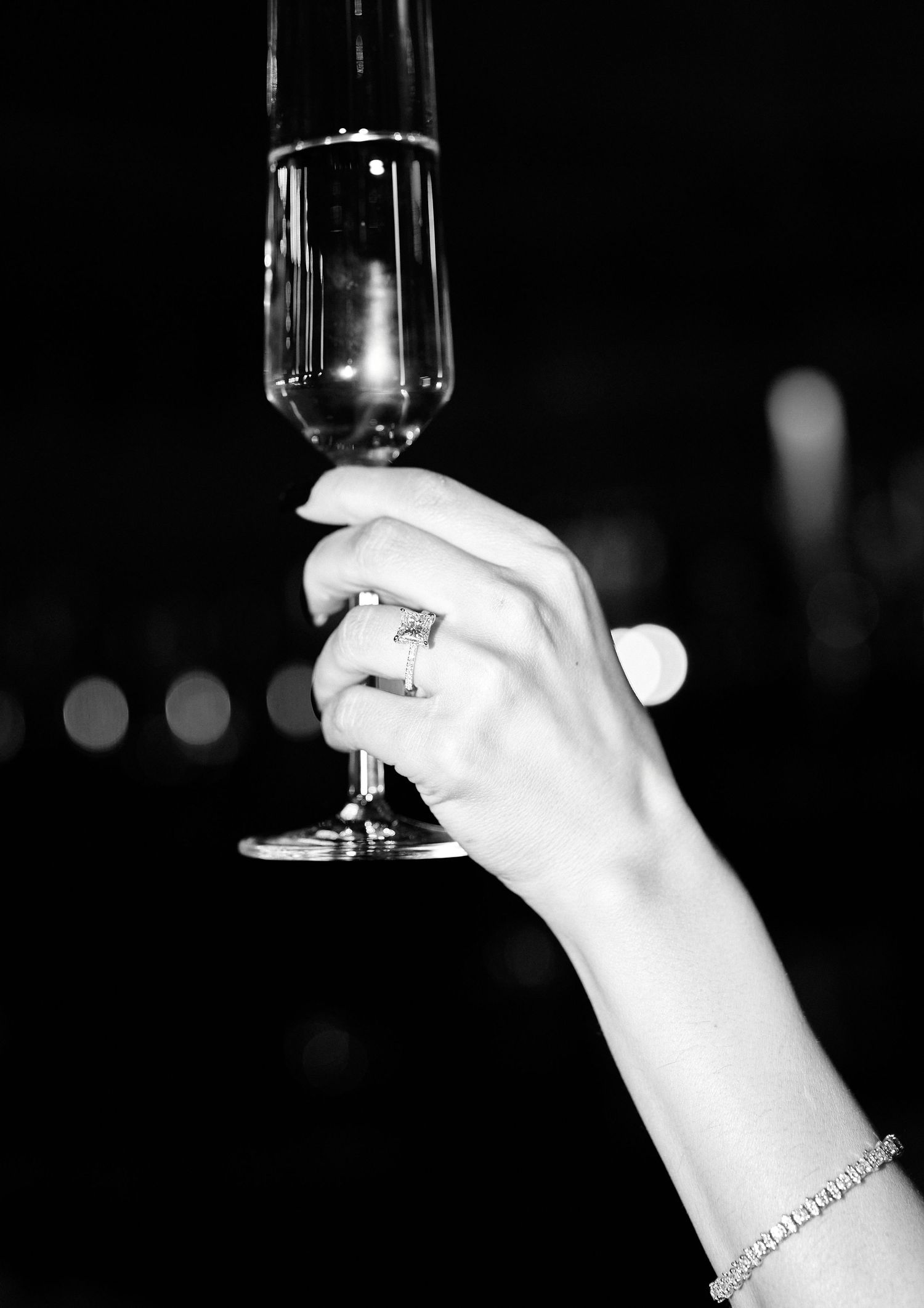 Black and white photo of a woman raising a champagne flute, wearing a diamond engagement ring and bracelet.
