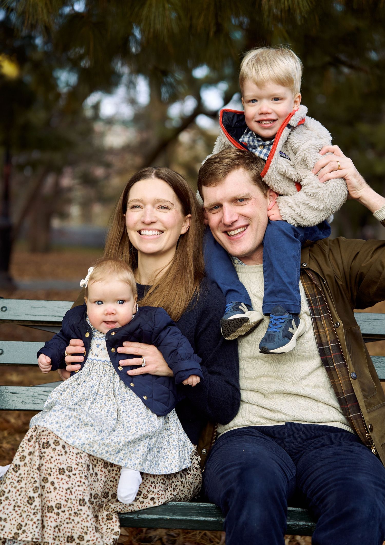 Happy family of four sitting on a park bench, parents smiling with toddler and baby in autumn.