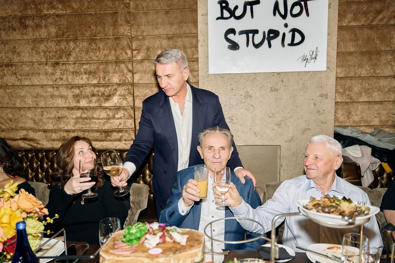 Guests raise glasses in a toast at an elegant dinner party beneath a bold 'But Not Stupid' sign.
