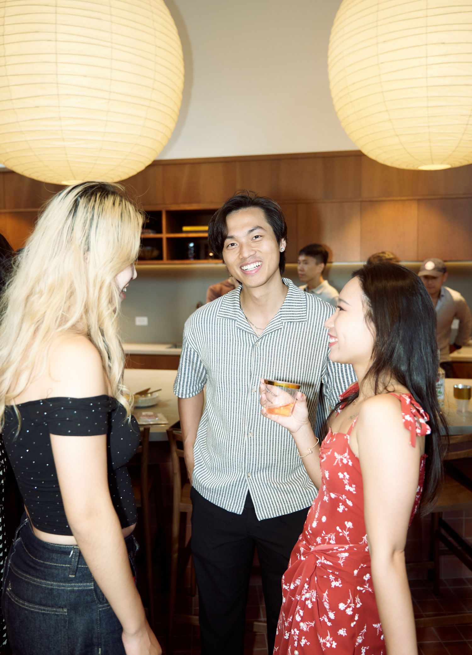 Three young adults laugh and chat while holding drinks under round paper lanterns at a social event.
