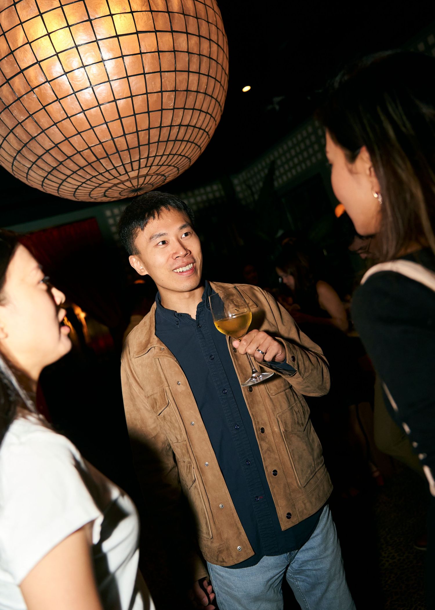 Man in brown jacket holds wine glass while conversing with guests at a dimly lit party venue.