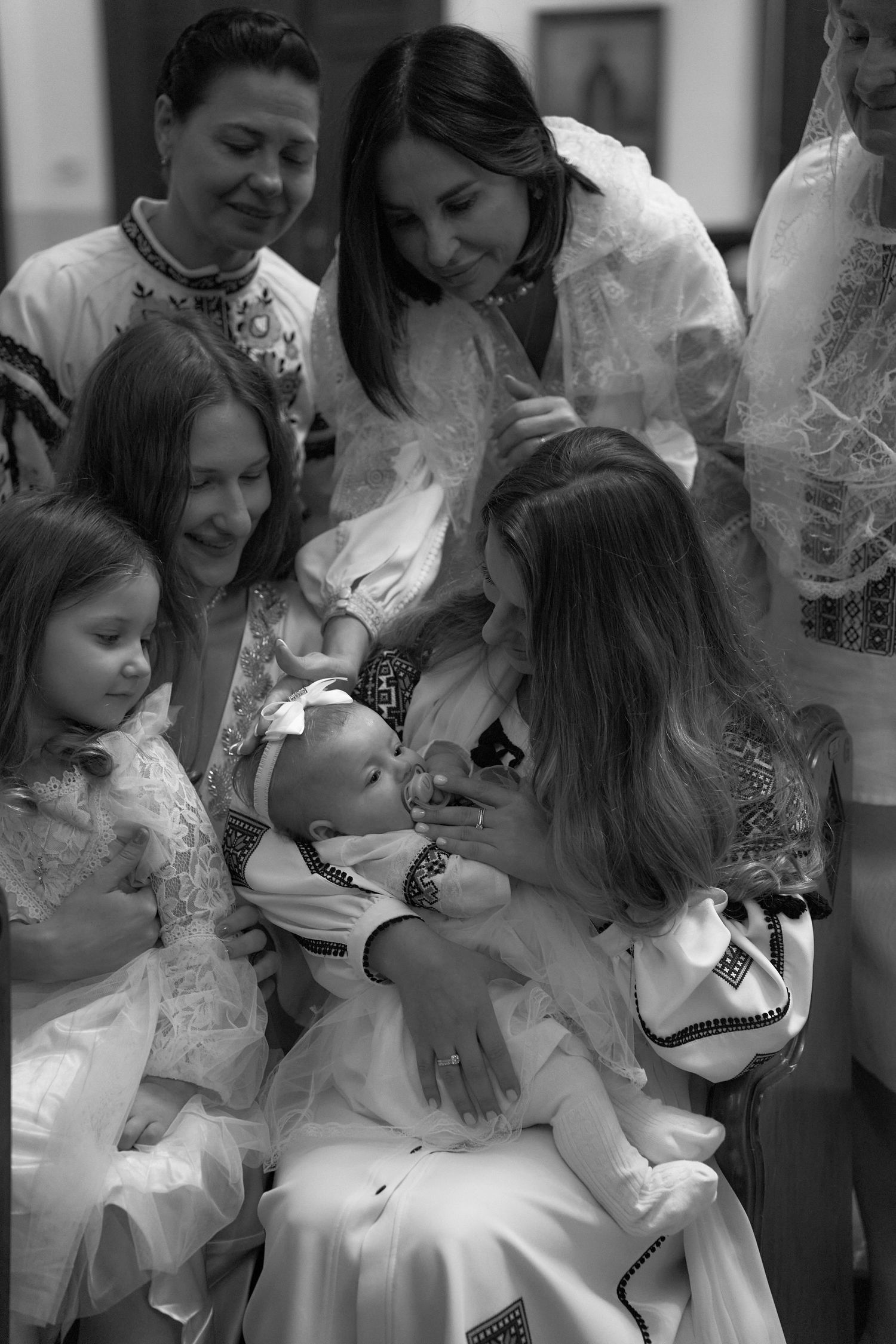 Black and white photo of women in embroidered white dresses gathered around newborn baby.